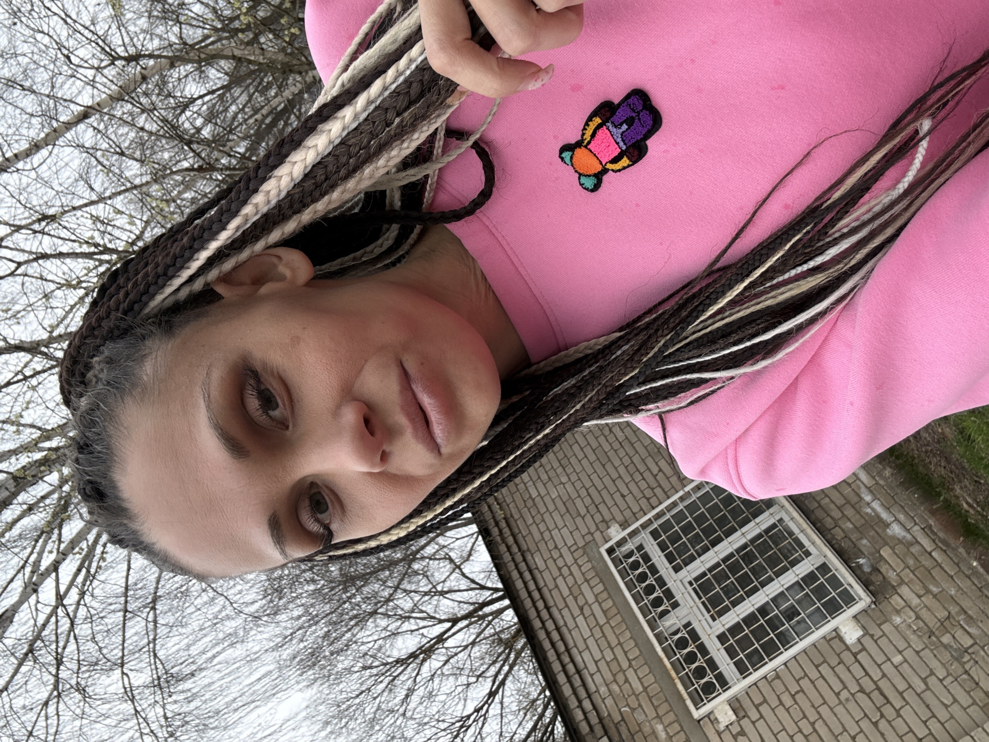 A selfie shows a woman with brown and white braids, wearing a pink sweater with a colorful butterfly patch. The background includes a grey brick structure with a window covered by a grid, along with bare tree branches. The location is Unknown, Unknown.