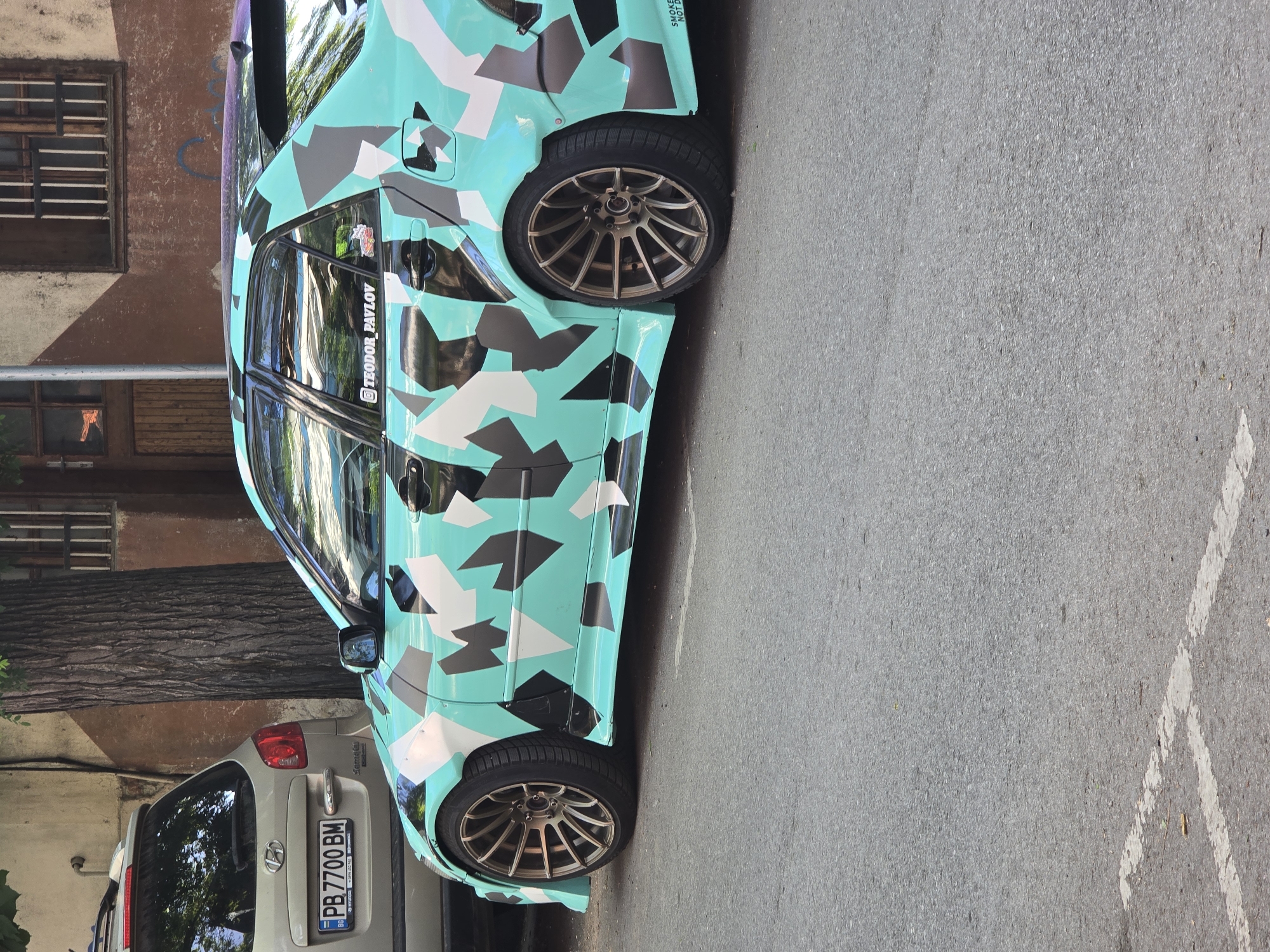 The image depicts a parked, customized car with a camouflage paint scheme in an urban setting. The car's base color is a light turquoise, overlaid with geometric shapes of black, gray, and white. The wheels are gold-colored with a multi-spoke design. A silver or gray car is parked behind it, with a visible license plate that reads "PB 7700 BM." The backdrop features a building with brown window frames and a portion of a tree trunk. The street is asphalt.