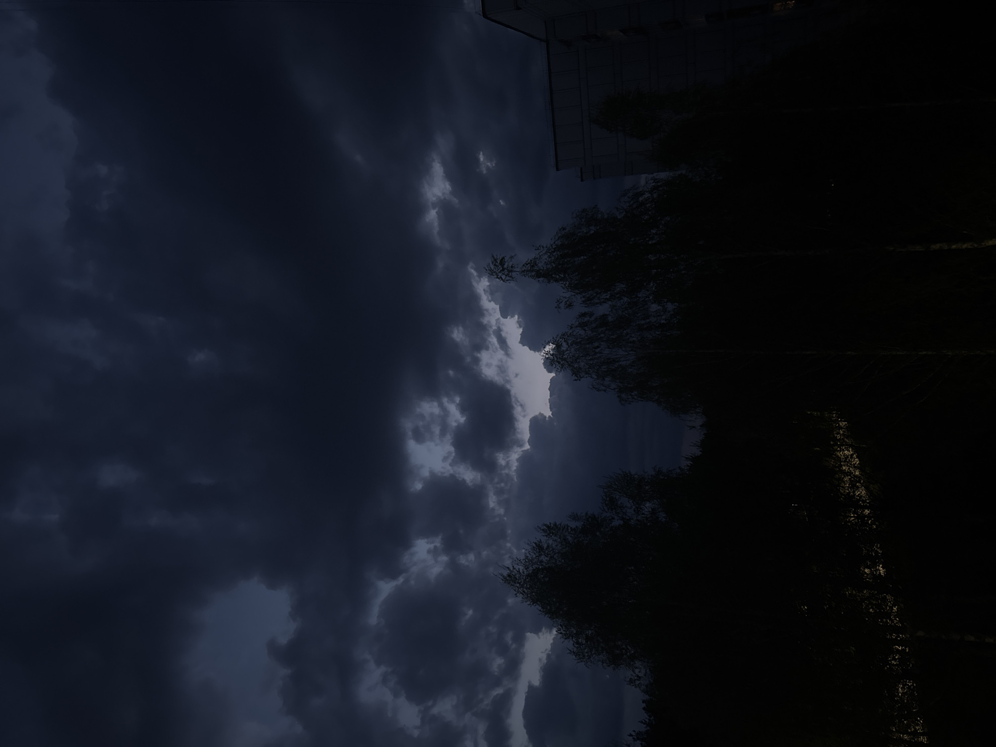 The image shows a night scene in what is presumed to be Chaikovsky, Russia. The sky is dominated by dark, stormy clouds. Some light is visible breaking through the clouds. Silhouetted trees are present on the right side. A building is partially visible on the upper right side. The ground is obscured in darkness.