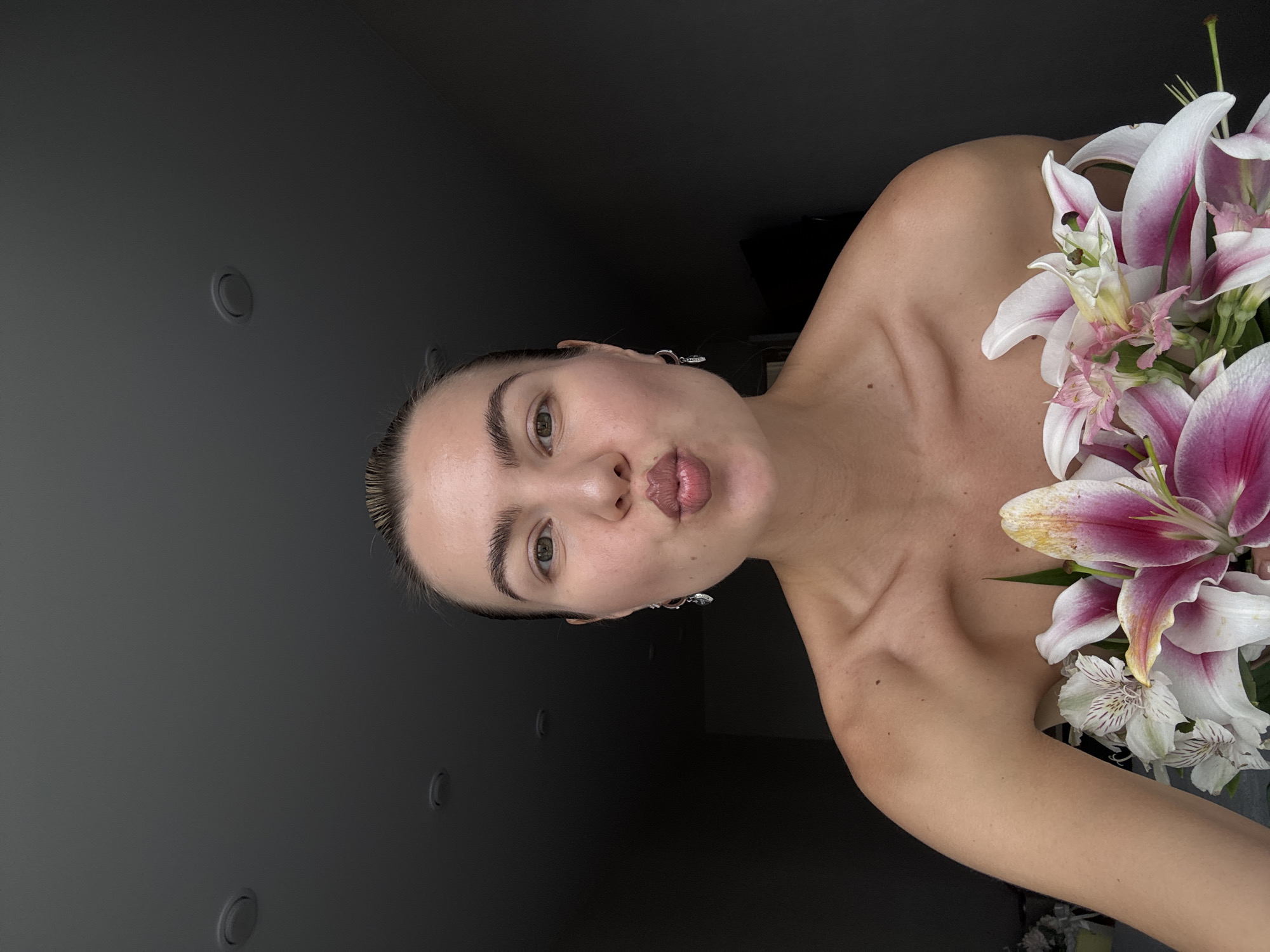 A woman with a bouquet of lilies is facing the camera and puckering her lips in a kiss. She has light brown hair slicked back, fair skin, and is wearing dangling earrings. The bouquet of lilies is positioned on her right shoulder, with large pink and white petals dominating the view. The background consists of two shades of gray, with a lighter gray wall on the left and a darker gray wall behind her. There are two circular recessed lights visible on the lighter gray wall. The location of this scene is Unknown, Unknown.