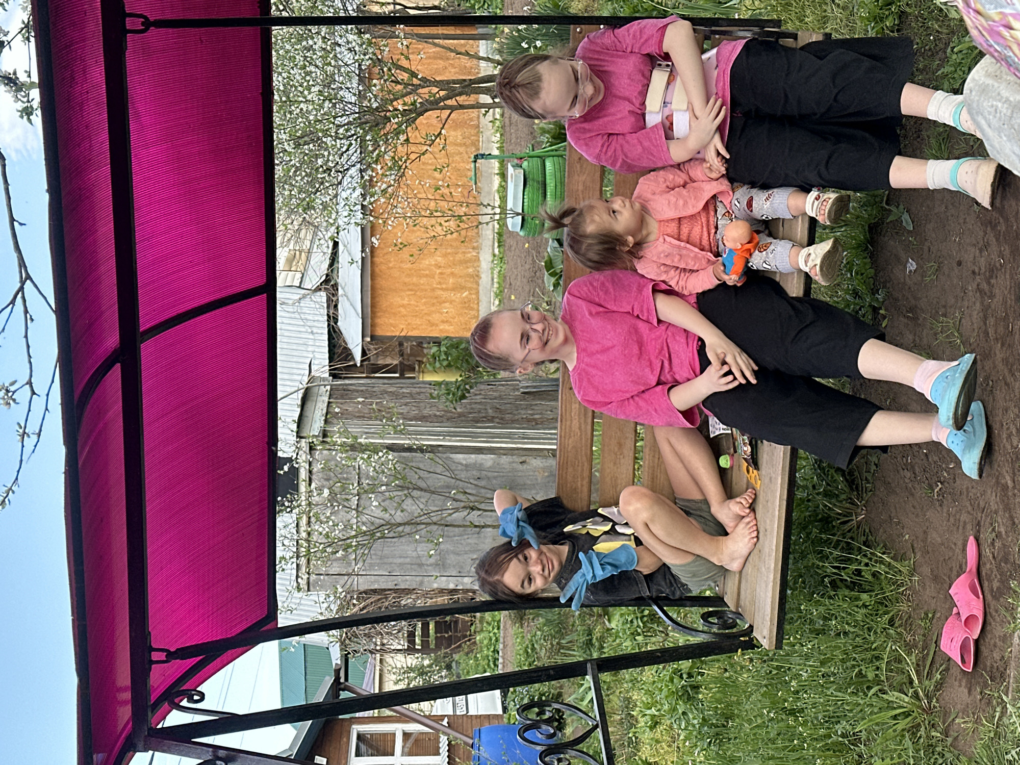 The image shows a group of four people sitting on a wooden swing in a backyard setting. The swing is covered by a pink canopy. Three individuals, including a woman, a girl, and a toddler, sit together on one side of the swing, all wearing pink tops. The woman wears glasses. The toddler holds a small doll. On the other side of the swing, a younger individual wearing blue gloves poses with hands raised. The environment includes a house, a wooden fence, green grass, and trees with white blossoms. A pair of pink sandals lies on the ground. The location is possibly Uva, Russia.