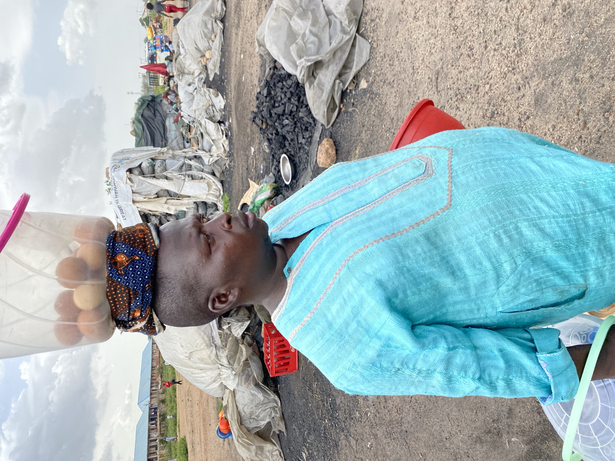 A person wearing a turquoise tunic stands in what appears to be an outdoor market or open area. The person has a bucket filled with eggs balanced on their head, secured by a patterned fabric. In the background, there are various piles of goods covered in tarps, some of which appear to be charcoal, and a few vehicles. Buildings with blue roofs are visible in the distance. The ground is unpaved and appears to be dirt and gravel. A red plastic bucket is visible near the person's hip. The scene is consistent with a street market environment in Bukuru, Nigeria.