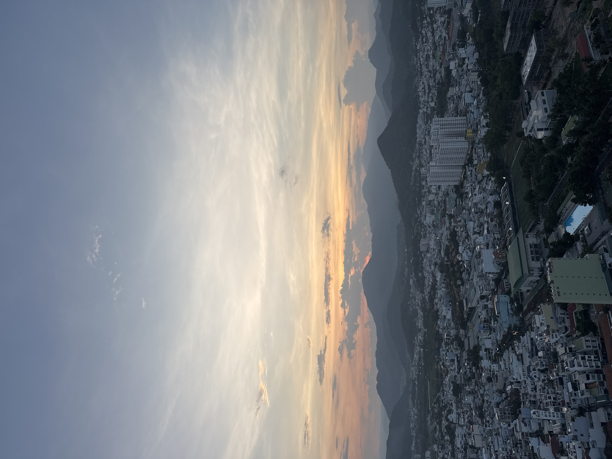 An aerial, high-angle, panoramic shot captures a city at dusk. The sky is a gradient of blue to pale orange and pink, with wisps of clouds, indicating the sun is setting behind the mountains in the distance. The city below is densely populated with buildings of varying heights and architectural styles, mostly in muted tones of white, grey, and light brown. Some buildings appear to be residential blocks, while others may be commercial or industrial. In the middle ground, there are several taller, modern buildings with many windows. Trees and greenery are interspersed throughout the city, and a dark mass of mountains forms a backdrop to the urban landscape. The overall impression is one of a sprawling city at the edge of a natural, mountainous region as daylight fades.