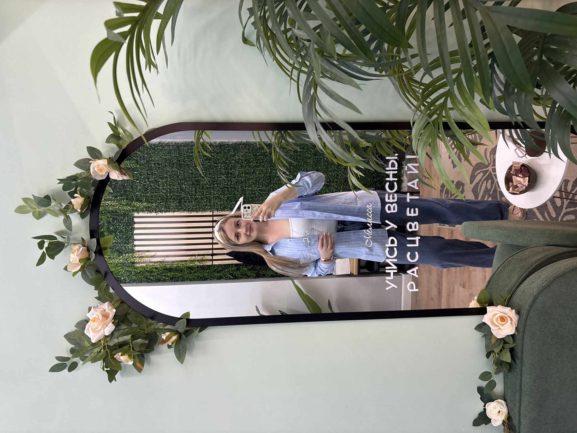 A woman stands in front of an arched mirror, holding a cell phone in her right hand and a plastic cup with a straw in her left. She is wearing a light blue striped shirt over a white tank top and blue jeans. The mirror is decorated with artificial pink roses and green leaves around its top edge. Reflected in the mirror is a woman with blonde hair, wearing a white headband, and a wall covered in artificial greenery with vertical wooden slats. White text is visible on the mirror below the reflection, reading "Мелисса УЧИСЬ У ВЕСНЫ, РАСЦВЕТАЙ!". To the left of the mirror is a pale green wall, and to the right, a large potted palm tree partially obstructs the view. In the foreground, a dark green upholstered chair is visible, adorned with two artificial white and pink roses. A small white table with a bowl of chocolates sits to the right of the chair. The floor is covered by a patterned rug. The scene suggests an indoor setting, possibly a cafe or a beauty salon, located in Chaikovsky, Russia.