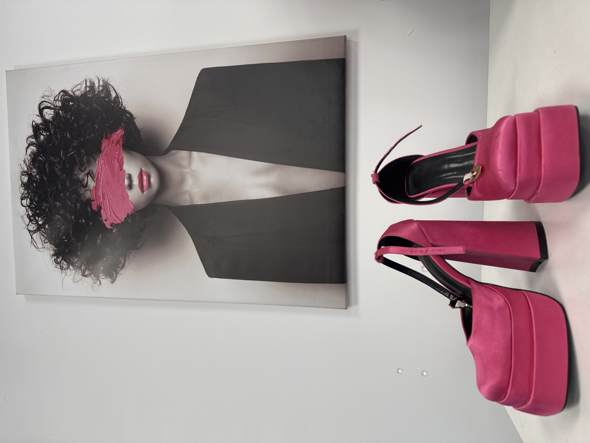 A photograph shows a portrait hanging on a white wall, next to a pair of bright pink platform shoes. The portrait is a stylized image of a woman with curly hair, partially obscured by a smear of pink paint. She wears a black blazer with a deep v-neck. The shoes, also bright pink, have thick platforms and straps. There is no indication of the location being Izhevsk, Russia, or any other specific city.