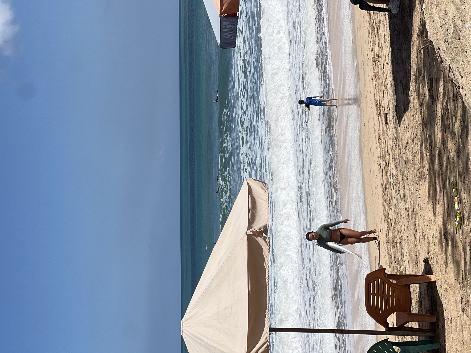 The image depicts a sunny day at a beach in Kuta, Indonesia. The scene shows a sandy beach with a gentle wave breaking on the shore. In the foreground, a light beige umbrella shades a part of the beach, and a brown plastic chair sits nearby, suggesting a relaxed atmosphere. A woman in a dark green long-sleeved wetsuit and black bikini bottoms is sitting on the sand, holding a white surfboard. She is looking towards the camera with a slight smile, indicating a positive emotion.
Further out in the turquoise water, a young boy in a blue shirt and shorts is walking near the shore, and another person is visible in the distance, possibly surfing. The sky is a clear, bright blue with a few wispy clouds. The presence of the umbrella, chair, and surfboard strongly suggests a day of recreational activity and leisure at the beach. The lighting and bright sky imply it is likely midday. The overall impression is one of a pleasant beach day, filled with activities like surfing and relaxation.