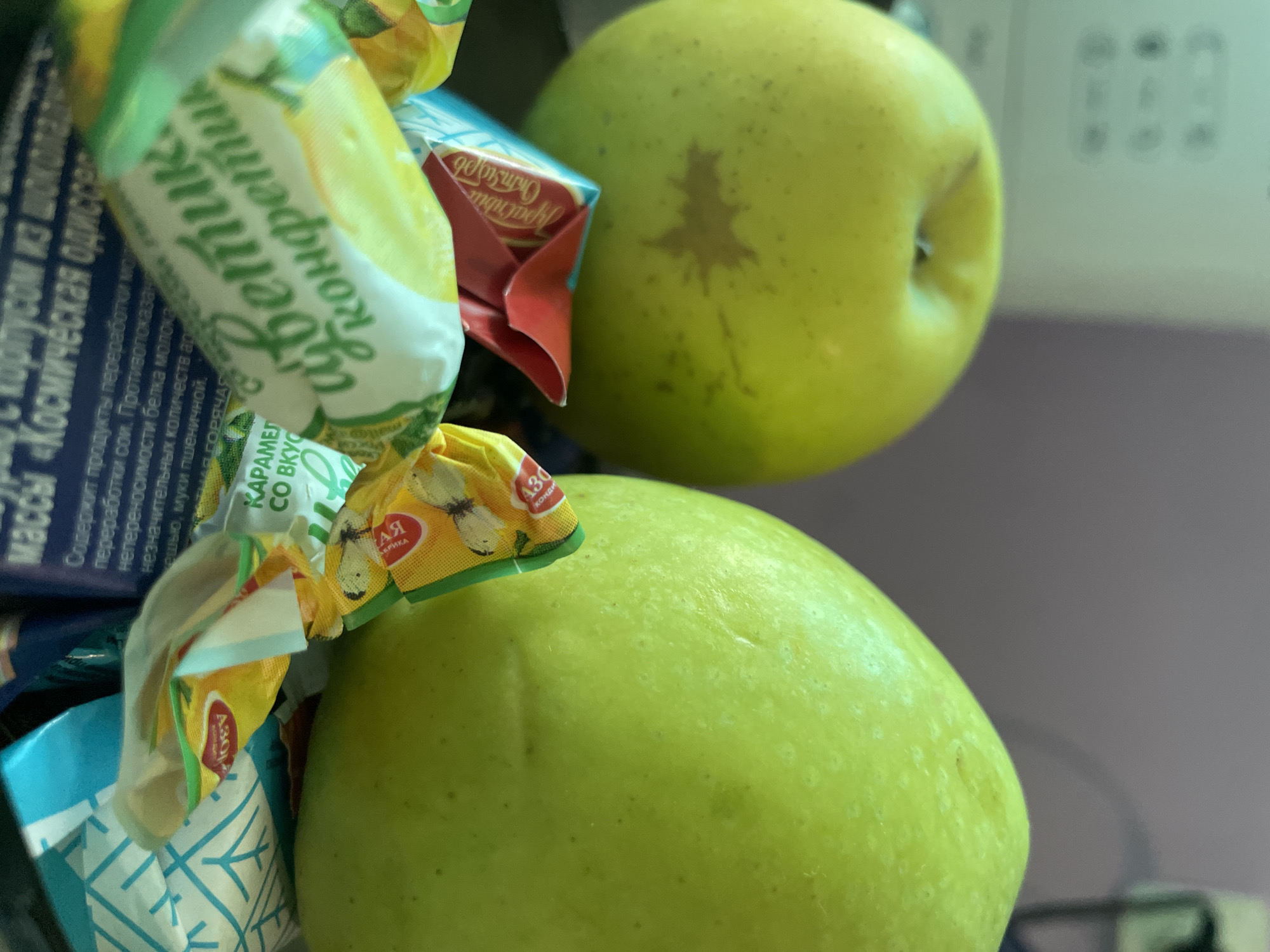 The image shows two green apples stacked on top of each other, surrounded by wrapped candies. The candies have Russian text on them, including the words "светик" and "АЯ" "Абрика". The apple on top has a darker tree-shaped mark on its skin. Part of a package with the words "массы «Космическая одиссе" can be seen. The background is blurred, but a part of an appliance can be seen. It is impossible to confirm if the photo was taken in Olginka, Russia.