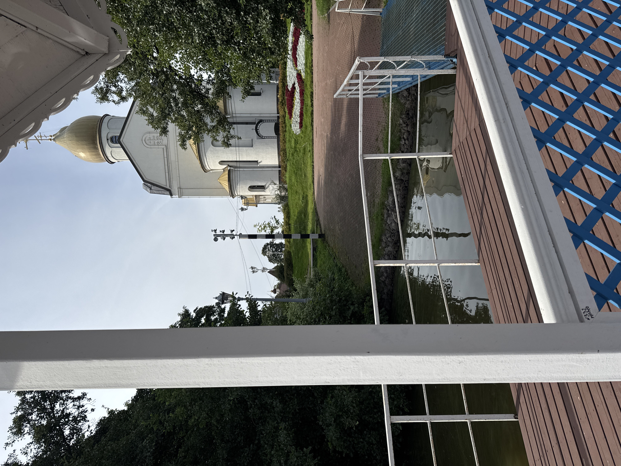 The image captures a view from a wooden pier overlooking a calm body of water, with a white Orthodox church featuring a golden dome in the background. The church is situated on a grassy hill, with flowerbeds in front of it. The pier has a white railing and a wooden deck. The scene is set in Sestroretsk, Russia, and appears to be a scenic and peaceful location. The sky is overcast, with a few clouds visible. The overall impression is one of tranquility and natural beauty, with the architectural grandeur of the church providing a focal point. There are no people visible in the image. The foreground of the image is dominated by the wooden pier and its railings. The water reflects the sky and the surrounding greenery. The church is the main subject in the background, with its distinctive golden dome and white facade. The landscaping around the church includes well-maintained grass and colorful flowerbeds.