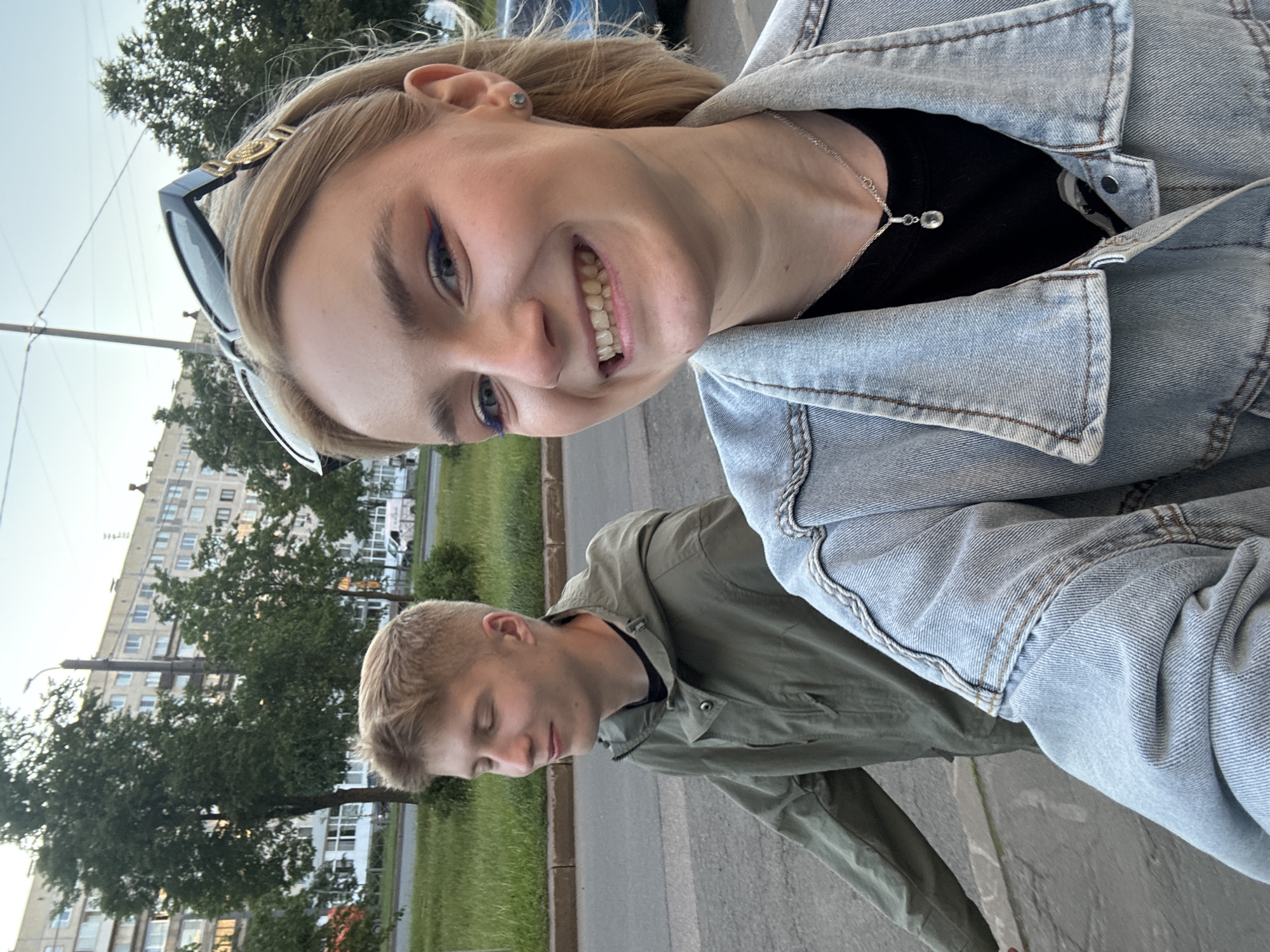 Two young people, a woman and a man, are captured in a selfie-style shot outdoors. The woman is in the foreground, smiling broadly and looking directly at the camera. She has blonde hair, is wearing makeup including blue eyeliner, and has on a denim jacket over a black top. She is also wearing sunglasses perched on her head. The man is in the background, slightly out of focus, with his eyes closed and a relaxed expression. He is wearing a green jacket. They appear to be standing on a sidewalk next to a grassy area. In the background, there is a multi-story apartment building and trees, suggesting an urban or suburban environment. The lighting indicates it is daytime, likely late afternoon or early evening given the soft light. The overall mood is casual and happy. The location is identified as Sinyavino, Russia. There is no visible text in the image.