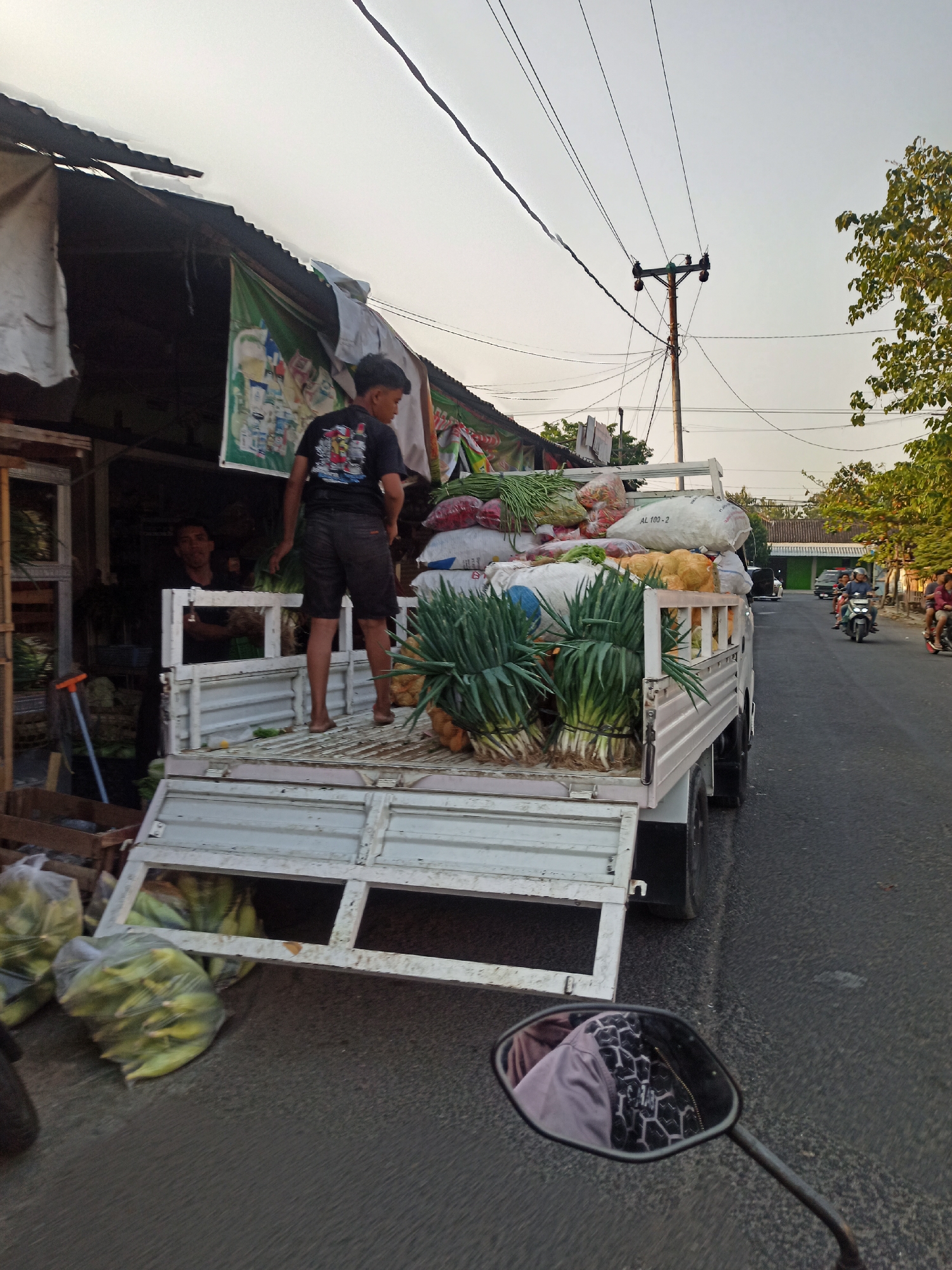 The image captures a street scene in Gemolong, Indonesia, likely a market area given the presence of a fruit and vegetable stall. A flatbed truck is loaded with fresh produce, including large bunches of green onions, bags of corn, and other vegetables. A young man in a black t-shirt and shorts stands on the back of the truck, appearing to be organizing or unloading the goods. Another man is visible inside the stall behind him. In the background, a utility pole with overhead wires stretches across the sky, and other vehicles, including motorcycles with riders, are visible on the road. The overall atmosphere suggests a bustling, early morning or late afternoon, typical of a market environment. The scene is shot from the perspective of someone riding a motorcycle, with the reflection of the rider and their clothing visible in the motorcycle's rearview mirror.
