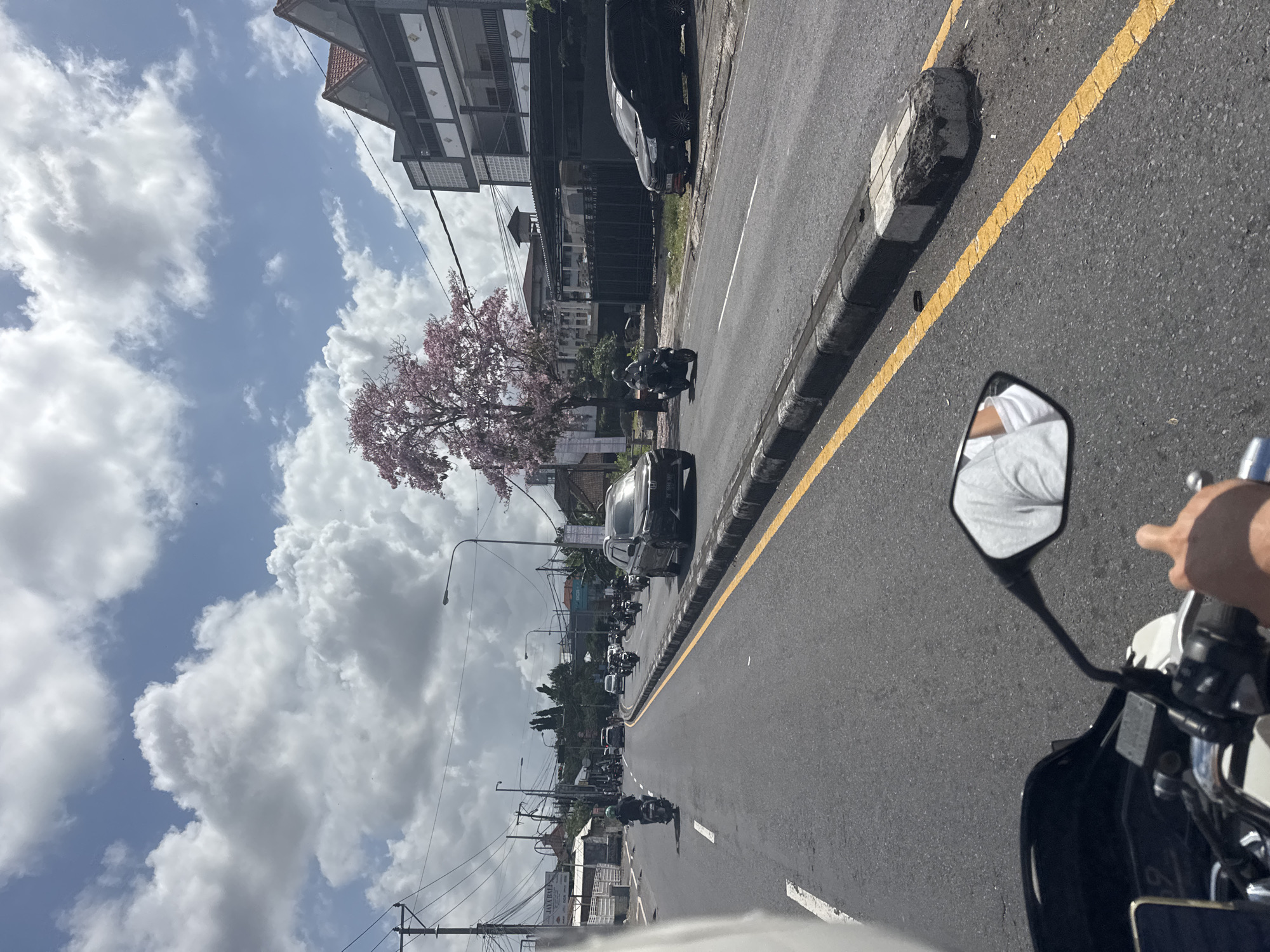 The image captures a street scene in Unknown, featuring a mix of buildings, trees, and vehicles. The sky is partly cloudy. A tree with pink blossoms is visible on the side of the road. Several cars and motorcycles are on the street. The perspective is from a moving motorcycle, with the rearview mirror and the rider's arm visible in the foreground.