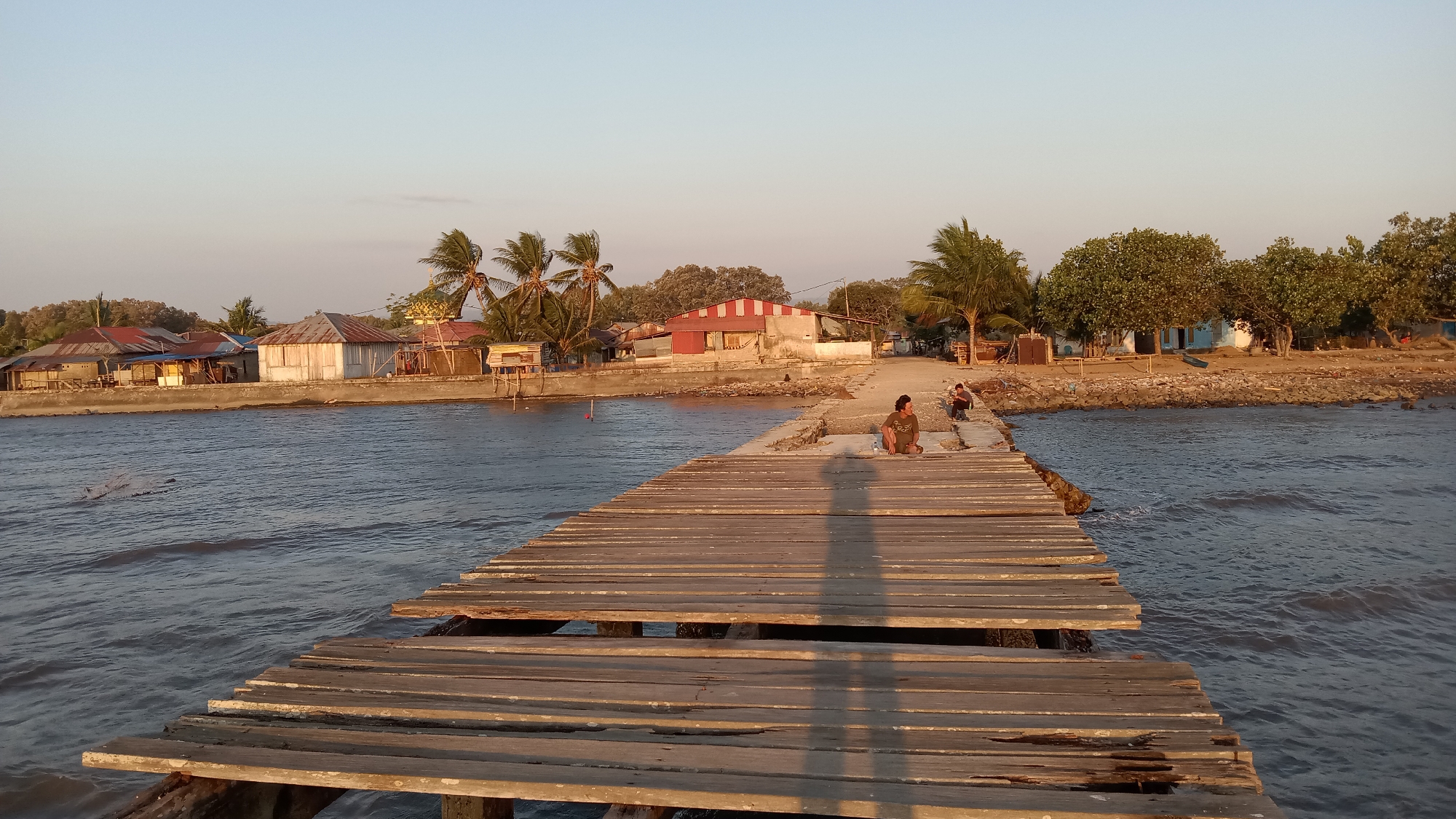 In the city of Unknown, Indonesia, a weathered wooden pier extends into the choppy waters of a body of water under a clear sky. The pier is in a state of disrepair, with several planks missing or broken. On the pier, two individuals are present. One person, dressed in a brown shirt, sits facing the water with their legs crossed, casting a long shadow onto the pier. Another person, also in dark clothing, is seated further down the pier, facing away from the camera. In the background, a coastline features a collection of buildings with red and silver corrugated roofs, typical of residential structures. Palm trees and other foliage dot the landscape, with the warm light of either sunrise or sunset casting long shadows and illuminating the scene. The water exhibits gentle waves, indicating a calm day.