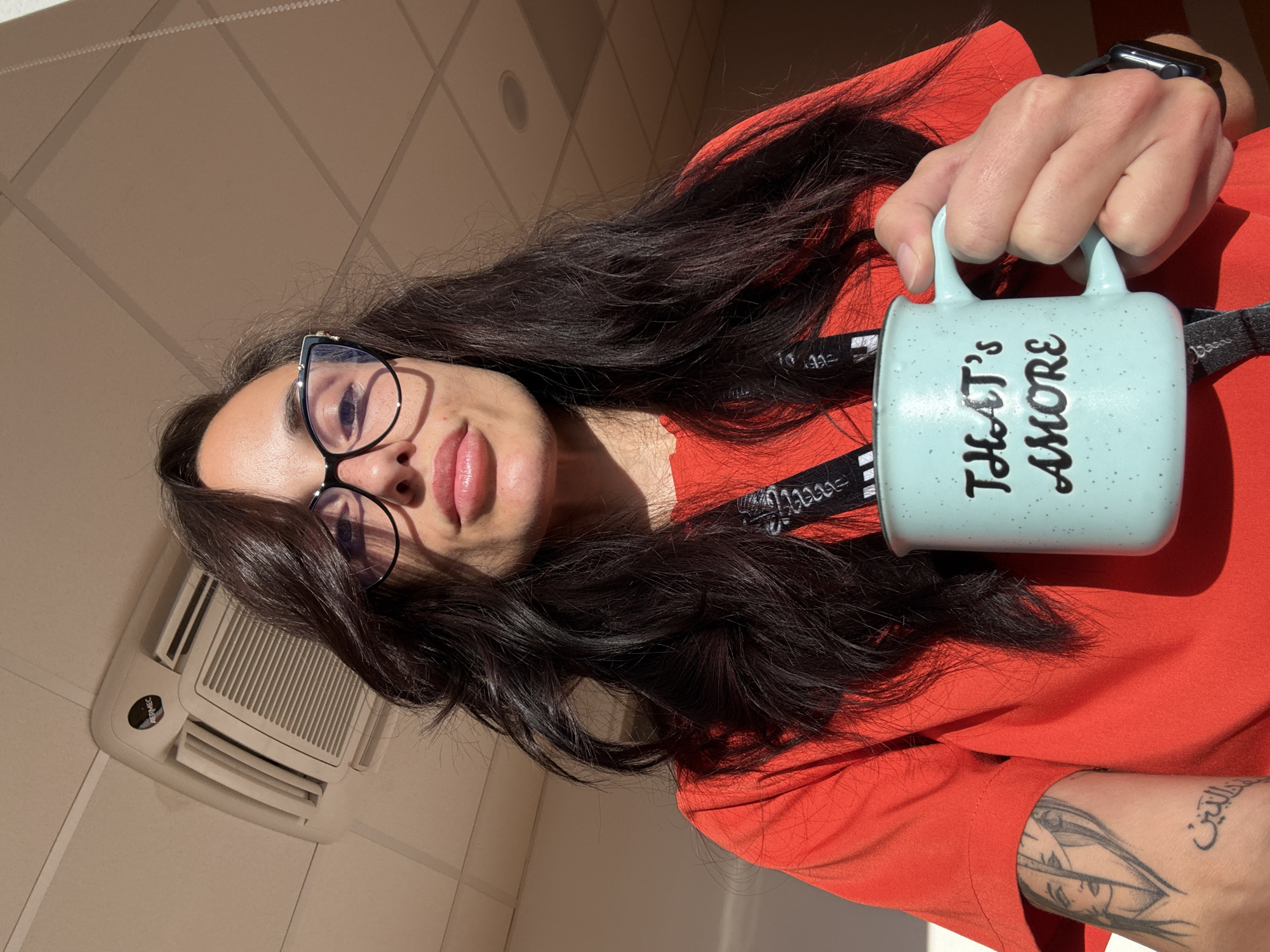 A woman is holding a light blue mug with the words "THAT'S AMORE" printed on it. She is wearing glasses and an orange shirt. She has dark hair and tattoos on her left arm. The location appears to be indoors, with an air conditioning unit visible on the wall. The location in the city of Pleven, Bulgaria, cannot be determined from the image.