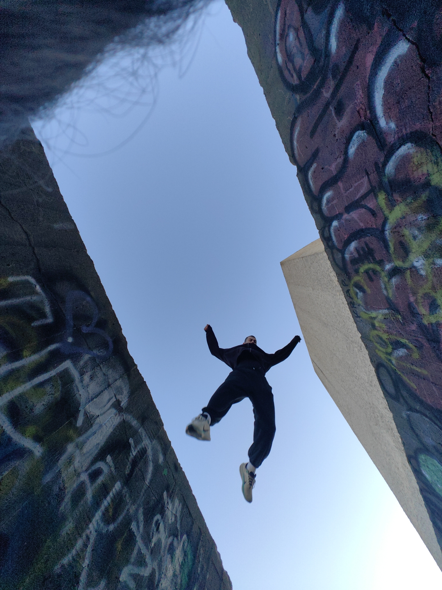 This low-angle photograph captures a dynamic moment of a person jumping against a clear blue sky, framed by two large, heavily graffitied concrete structures. The individual, appearing young and dressed in dark clothing (black hoodie and pants) and light-colored sneakers, is suspended in mid-air with arms slightly raised and legs bent, suggesting an energetic leap or fall.
The setting appears to be an urban or industrial outdoor space, characterized by the substantial concrete walls that dominate the foreground and sides of the frame. These structures are extensively covered in vibrant, multi-colored graffiti, featuring various tags, abstract shapes, and lines. Dominant colors include shades of purple, green, black, white, and blue. On the left concrete structure, parts of what appear to be the word "REAL" and a faint blue heart shape are visible among other abstract tags and designs. The right structure also displays a complex mix of colorful spray paint.
The time of day is clearly daytime, indicated by the bright, cloudless blue sky and consistent lighting, which suggests fair weather. The low-angle composition emphasizes the height of the structures and the airborne subject, creating a sense of scale and freedom. While the person's facial expression is not clearly visible, the act of jumping conveys a sense of exhilaration, energy, or artistic expression. This scene is located in Varna, Bulgaria.