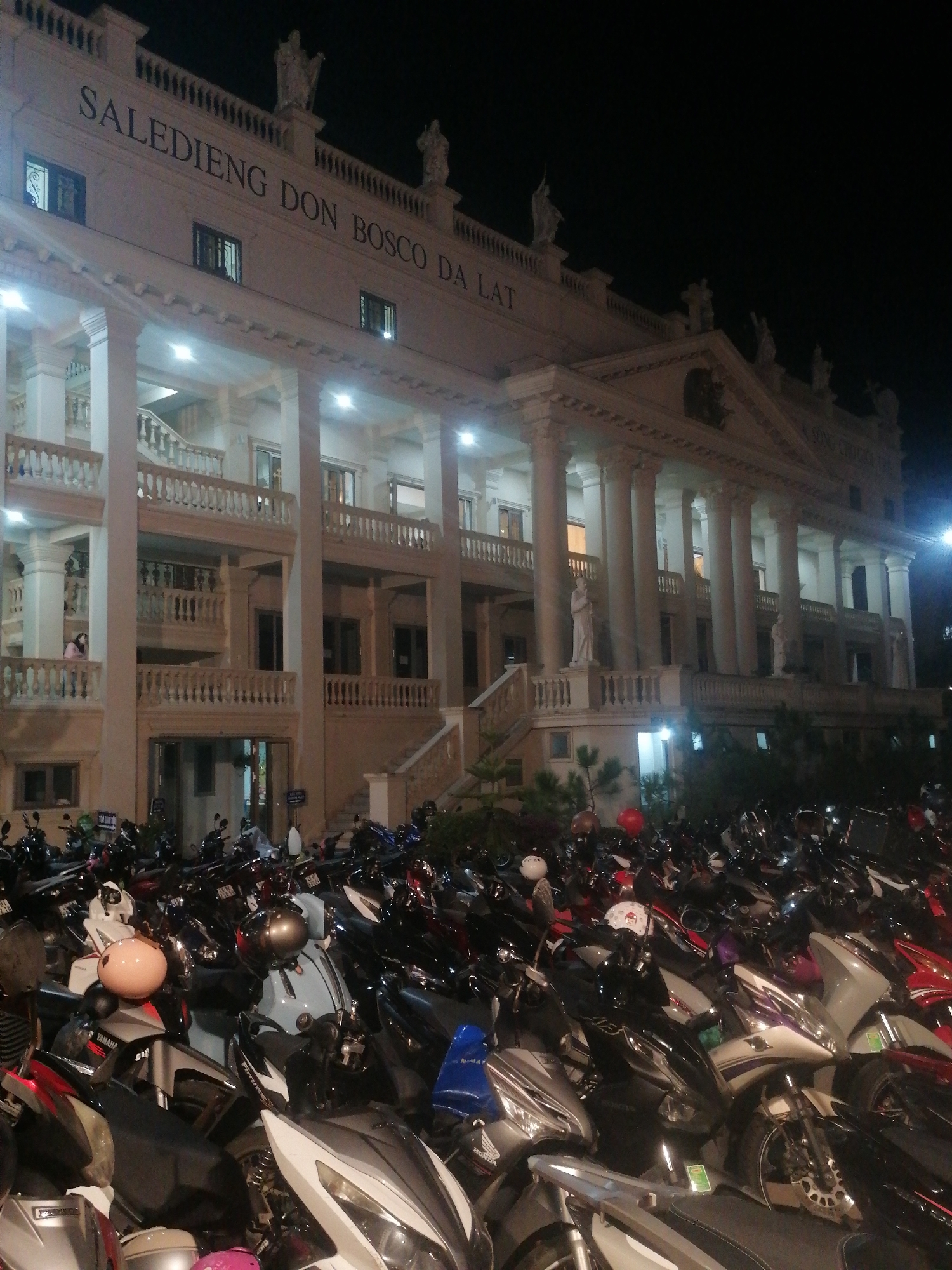 The image captures a nighttime view of the Salle dieng Don Bosco Da Lat, a large, multi-story building with a classical architectural style, characterized by numerous columns, balconies, and decorative statues. The building is brightly lit from within, illuminating the facade and casting light onto the surrounding area. In front of the building, a large number of parked motorcycles are visible, filling the foreground and extending into the mid-ground. The motorcycles are of various models and colors, and some have helmets placed on them. The scene appears to be a public space, possibly a parking lot or a gathering area. The presence of the motorcycles suggests it may be evening or nighttime, and a significant number of people are likely present or have recently been present at the location. The sky is dark, indicating it is night.