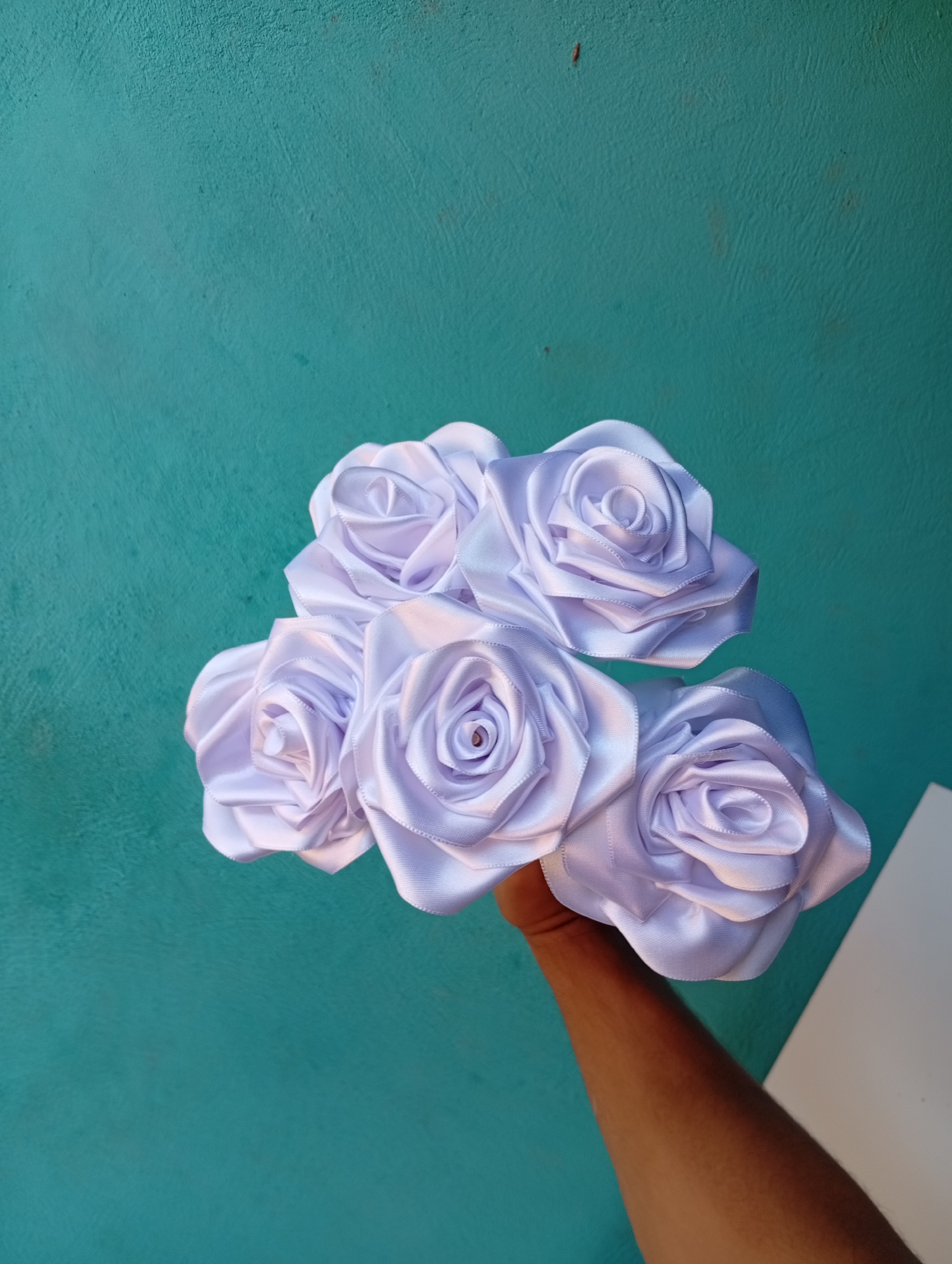 A person's arm and hand are holding a bouquet of five white satin roses against a teal-colored textured wall. The roses are arranged in a cluster, with three in the front and two behind them. The wall appears to be an exterior surface. The scene is photographed from a slightly low angle.