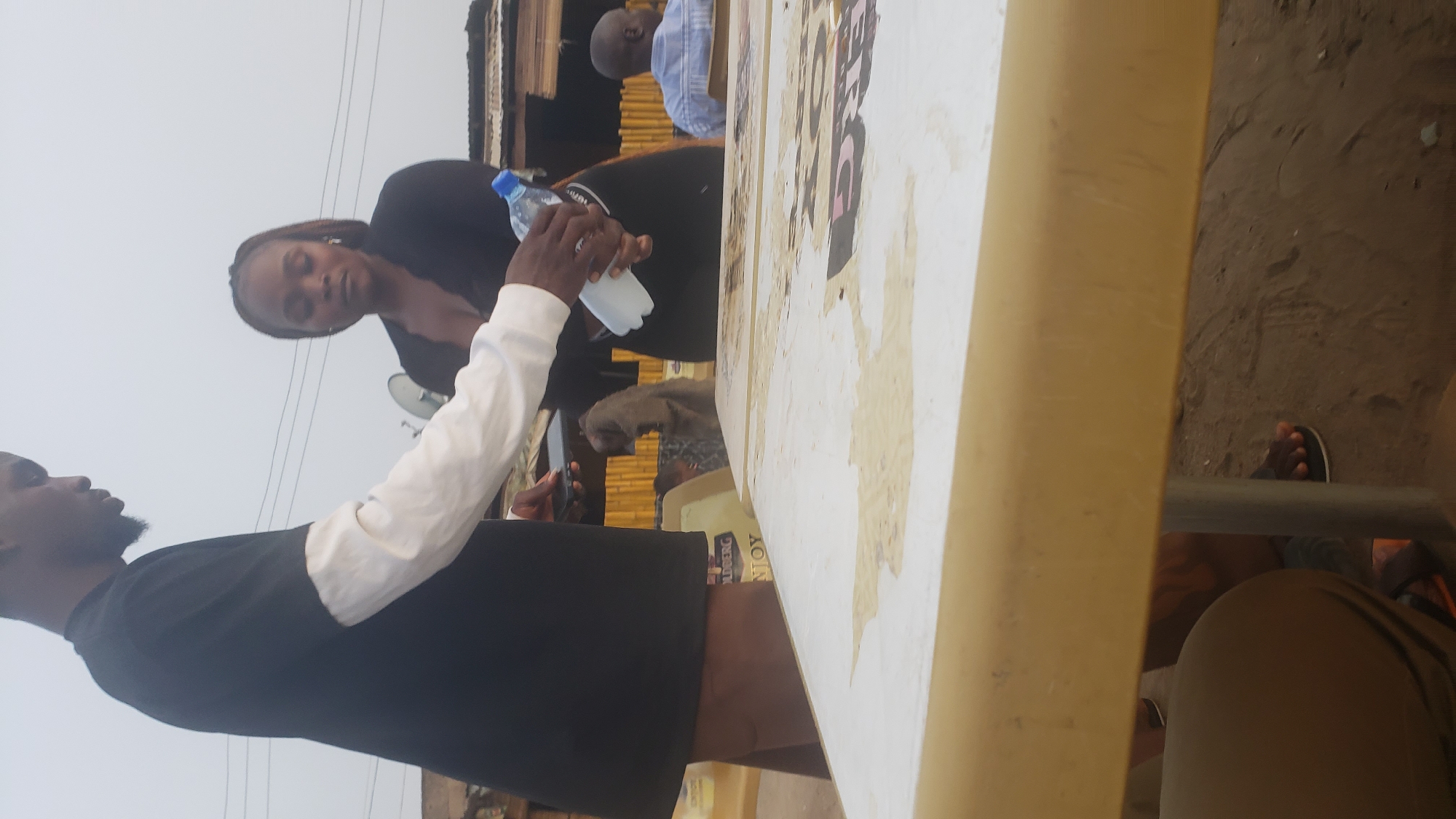 A medium, wide-angle shot depicts an outdoor scene in Narayi, Nigeria. A light beige table with peeling stickers dominates the foreground and right side of the frame.
A dark-skinned man in a black shirt with long white sleeves stands to the left. He holds a clear water bottle with a blue cap. Behind him, a dark-skinned woman in a black top looks down, perhaps at the water bottle or table. An antenna is behind her head.
In the background, another man in a blue shirt is partially visible. There are also several undefined people standing and sitting. The environment includes a brown, dusty ground. There are bamboo structures visible in the background.
A foot wearing a sandal can be seen under the table, at the bottom right corner of the frame.