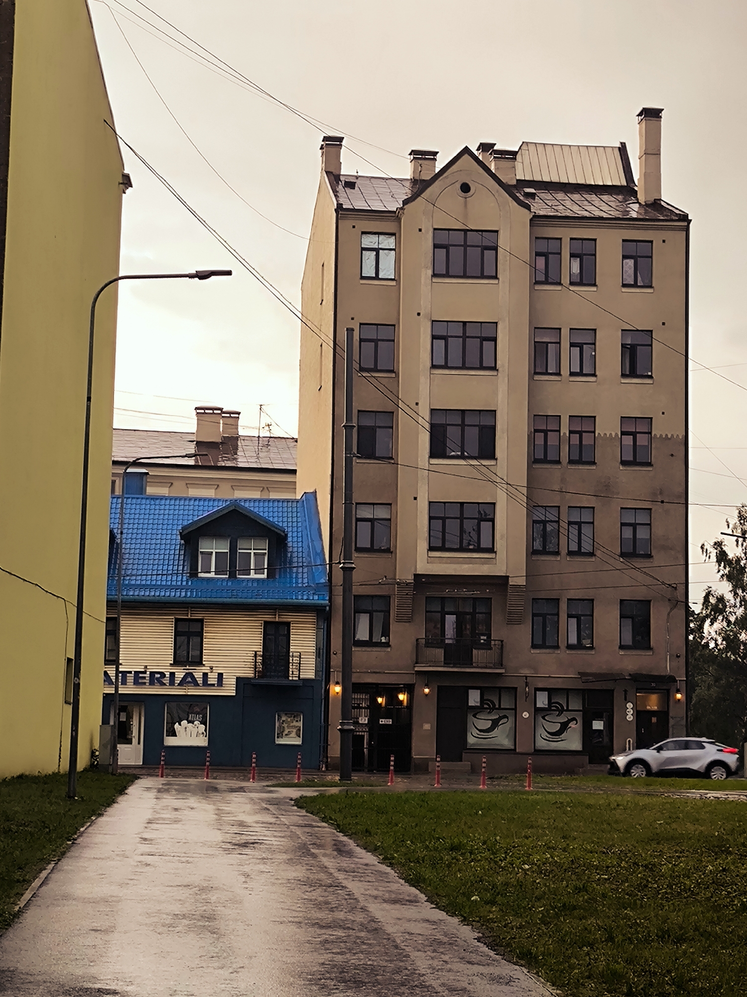 This image depicts a street scene in Riga, Latvia, likely in the evening or on an overcast day, judging by the lighting. On the left, a tall, smooth, pale yellow building dominates the foreground. To its right, a narrower, multi-story building with a distinctive bright blue tiled roof stands out. This blue-roofed building has a light-colored facade with dark windows and a balcony on the upper level. The words "MATERIIALI" are visible on the facade of this blue-roofed building.
Further to the right, a taller, light brown, multi-story building with numerous dark windows and visible chimneys rises. The architecture of this building appears older, possibly Art Nouveau or a similar early 20th-century style. There are overhead wires strung across the scene, typical of urban environments.
In the foreground, a wet, paved walkway or road curves towards the right. Adjacent to the walkway is a strip of green grass. A silver car is parked on the right side of the street, in front of the light brown building, which appears to house shops or businesses with cafe-style window decorations featuring coffee cups. Several orange traffic cones are placed along the edge of the walkway. The sky is a muted, light gray, suggesting cloudy weather.