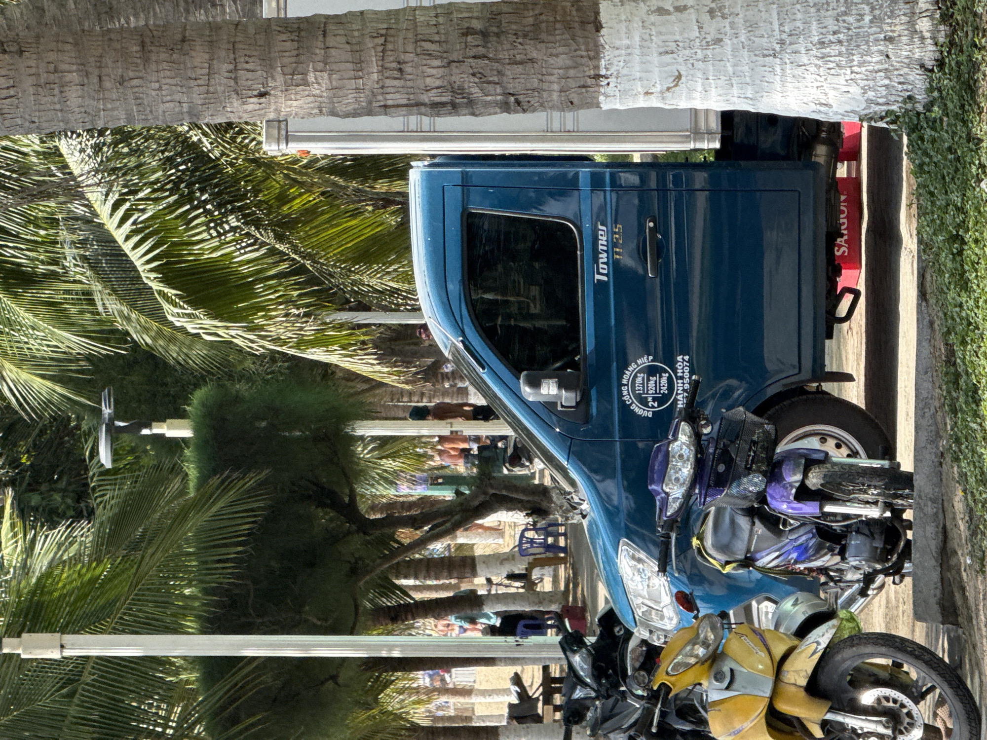 The image shows a blue Towner TT 25 truck parked on a street in Nha Trang, Vietnam, likely near a beach or park, as suggested by the palm trees and sand visible in the background. The truck is flanked by several motorcycles, including a yellow scooter on the left and a purple scooter in the middle. The truck's door has text including "Townel" and "TT 2.5", and what appears to be Vietnamese text and weight specifications like "1370kg" and "920kg". The truck has a white boxy cargo area. In the background, there are more palm trees, streetlights, and people, some of whom appear to be wearing beachwear. A red object with the word "SAIGON" is visible under the truck. The overall impression is of a warm, sunny day in a coastal city.