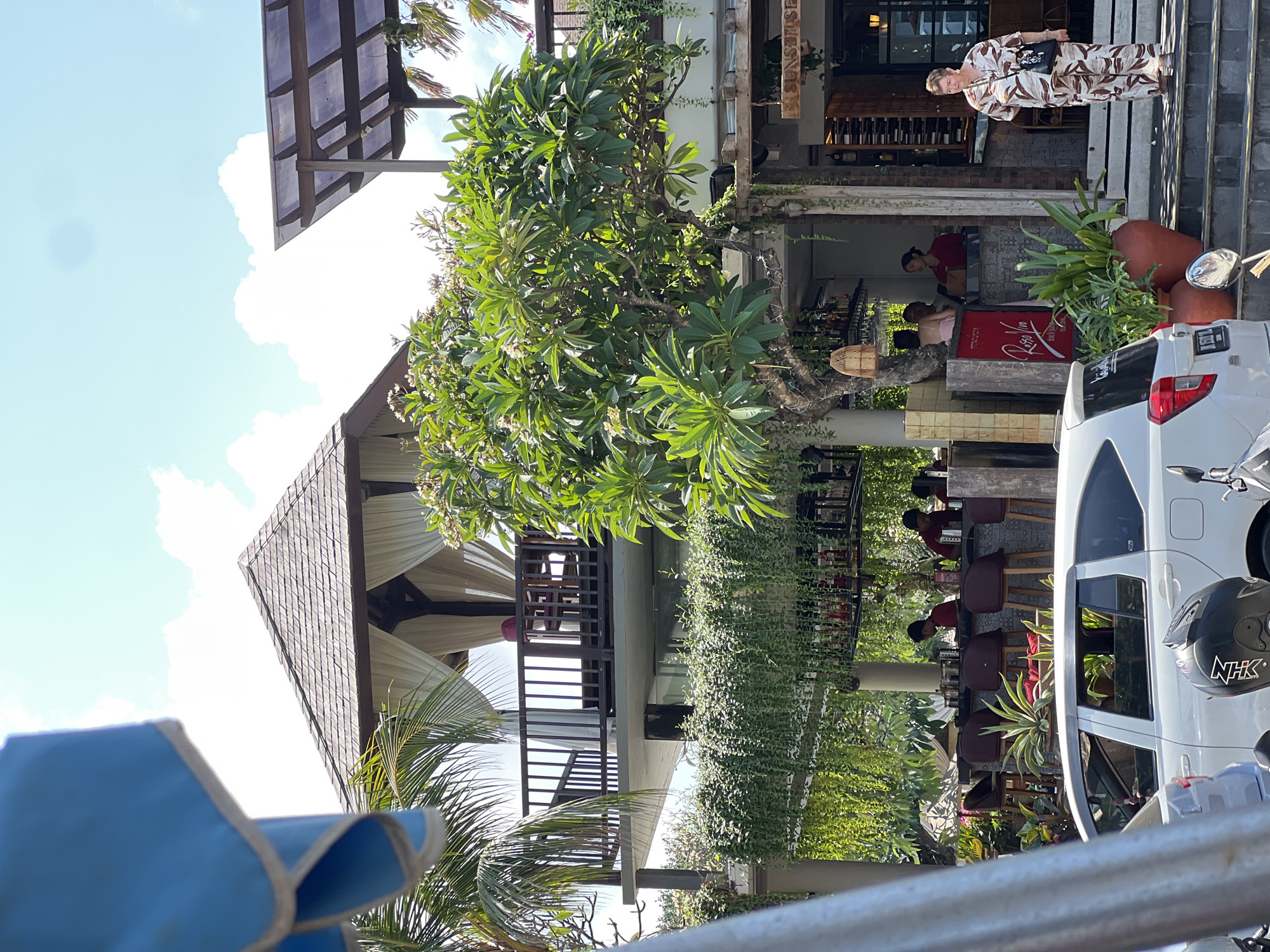 In Kuta, Indonesia, a woman in a patterned jumpsuit stands on steps leading up to an establishment with a sign reading "SUNSET BAR". Beside her, a white car is parked, and a black motorcycle helmet rests on its seat. In the background, a bar area is visible with several people sitting on stools, some appearing to be staff. Lush greenery hangs from the structures, and a large tree with broad leaves stands prominently. Above, a wooden-roofed structure with white curtains and a balcony suggests a multi-level building. The sky is bright with scattered clouds.