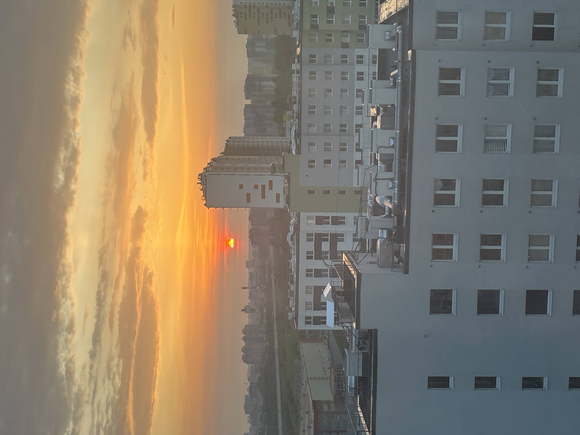 The image shows a cityscape at sunset, possibly in Kokkorevo, Russia. The sky is filled with orange and yellow clouds, and the sun is setting in the distance. In the foreground are several tall buildings, with many windows and white facades. The buildings are densely packed together, suggesting a densely populated urban area. Below the buildings is a hazy view of what appears to be a river or a long, straight road. There are no visible people or actions. The environment is hazy, likely due to pollution or the atmosphere at sunset.