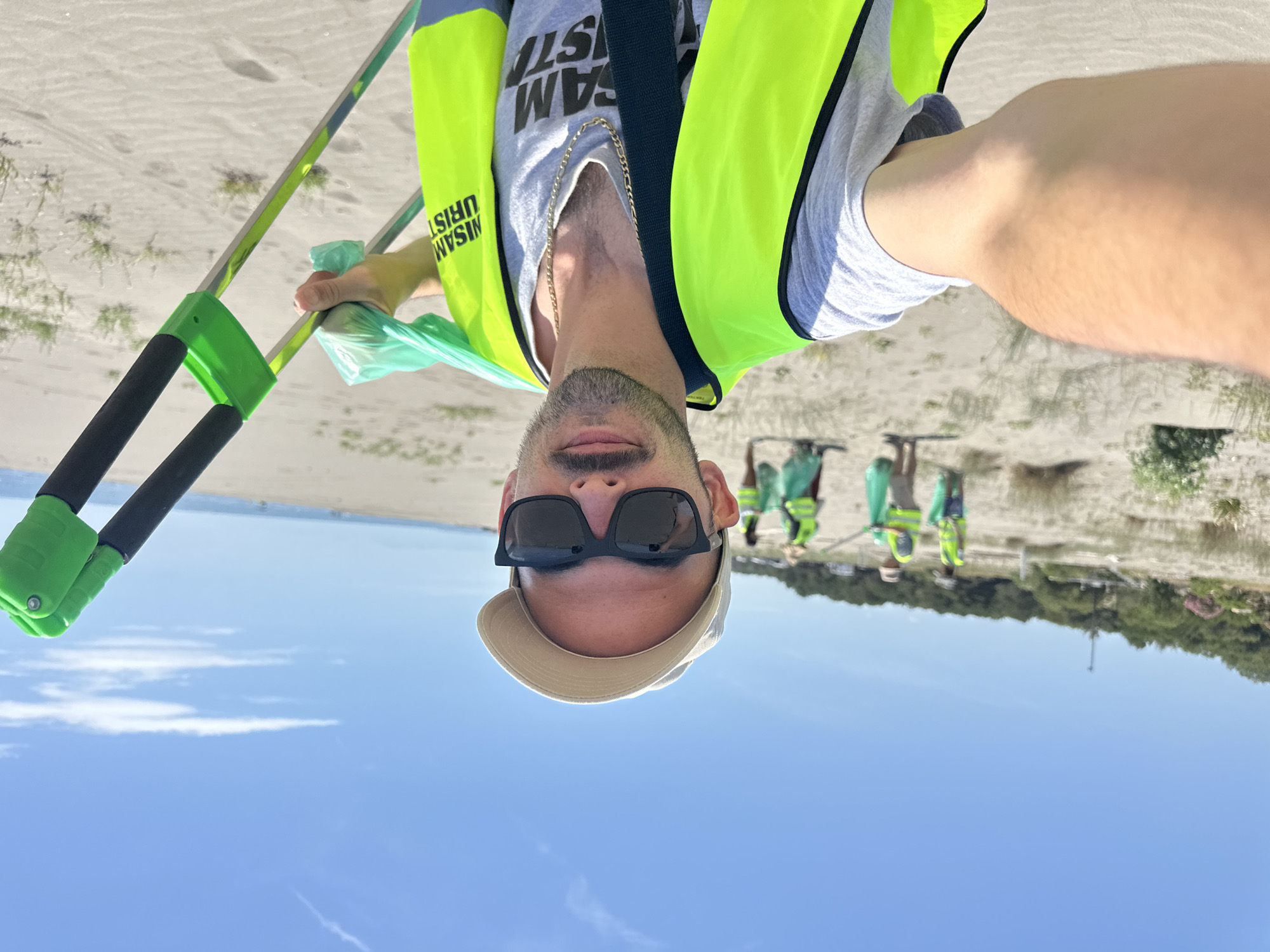 A selfie shows a man wearing sunglasses, a light-colored baseball cap, and a neon yellow safety vest over a grey t-shirt with black text, standing on a beach in Donji Štoj, Montenegro. He is holding a green and black trash picker in his left hand. A group of people wearing similar safety vests are picking up trash in the background. The beach has sand, some vegetation, and in the distance, some trees. The sky is blue. The image is rotated approximately 180 degrees.