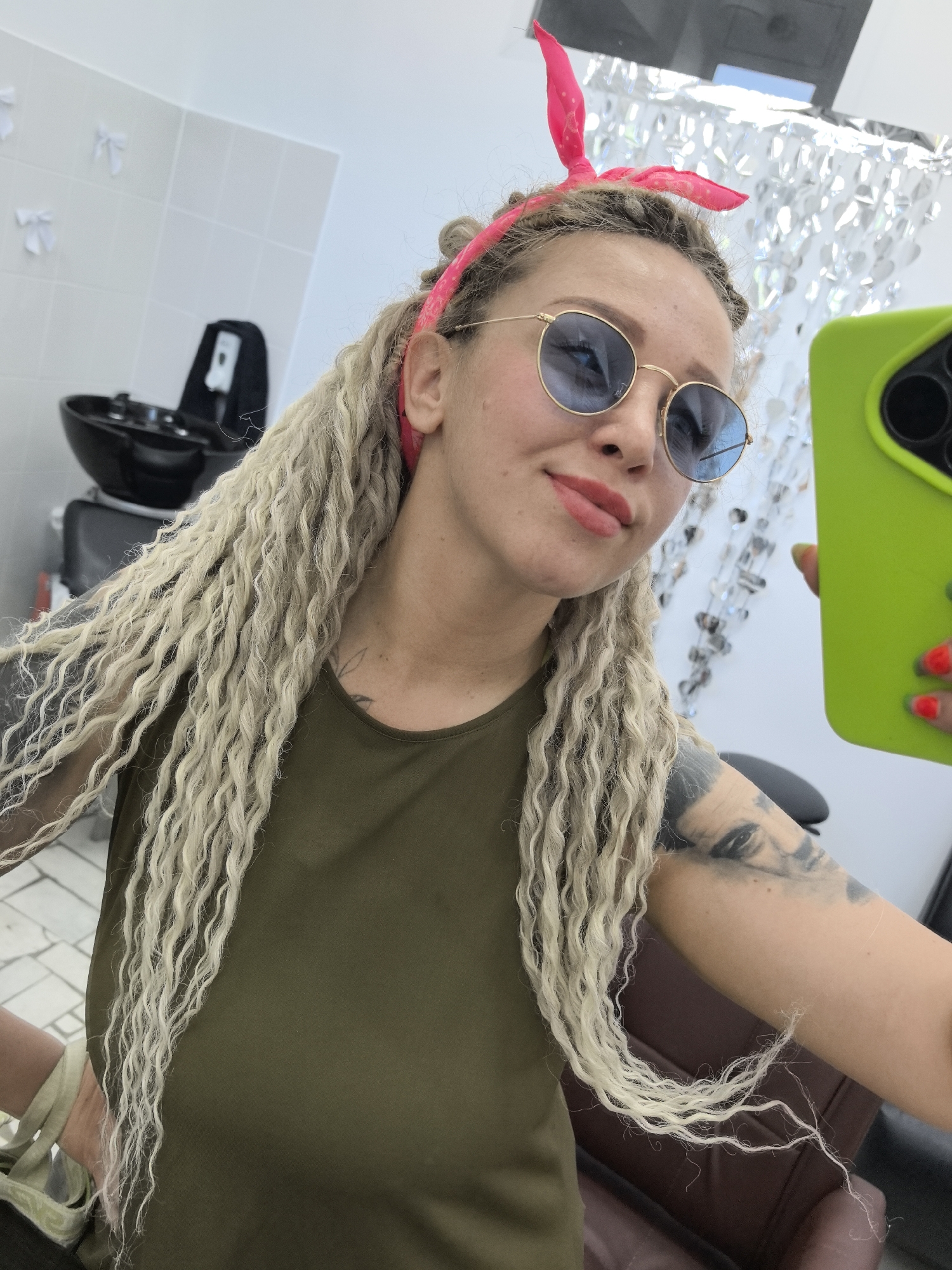 A woman is taking a selfie in what appears to be a hair salon. She is wearing a pink headband, sunglasses with blue lenses, and an olive green top. Her hair is styled in long, light-colored braids. Tattoos are visible on her arms. She is holding a bright green phone. In the background, there is a black hair washing station, white tiled walls decorated with white bows, and a silver, foil-like decoration hanging from the ceiling. There is no direct indication that this is the city of Chaikovsky, Russia.