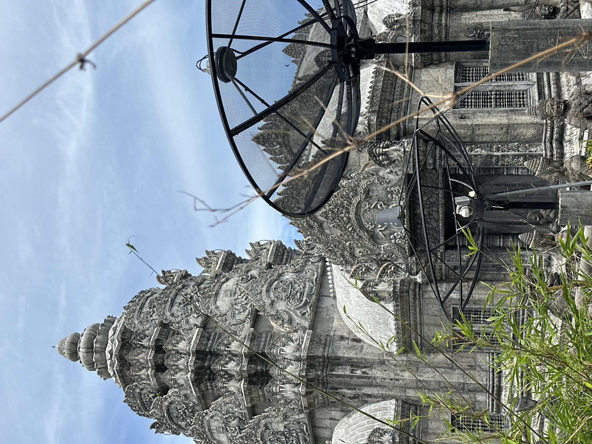 The image captures a weathered, intricately carved stone temple structure in the Ko Pha-ngan District of Thailand, with a large black satellite dish prominently positioned in the foreground, partially obscuring the temple's architecture. The temple features multiple tiered roofs adorned with detailed sculptures of mythical figures and serpents. Several stone statues of what appear to be seated guardians are visible at the base of the structure. Lush green bamboo shoots and dry, thin branches in the foreground add a natural element to the scene, creating a contrast with the man-made structures. The sky above is a mix of blue with wispy white clouds. The lighting suggests it is daytime, possibly late morning or early afternoon.