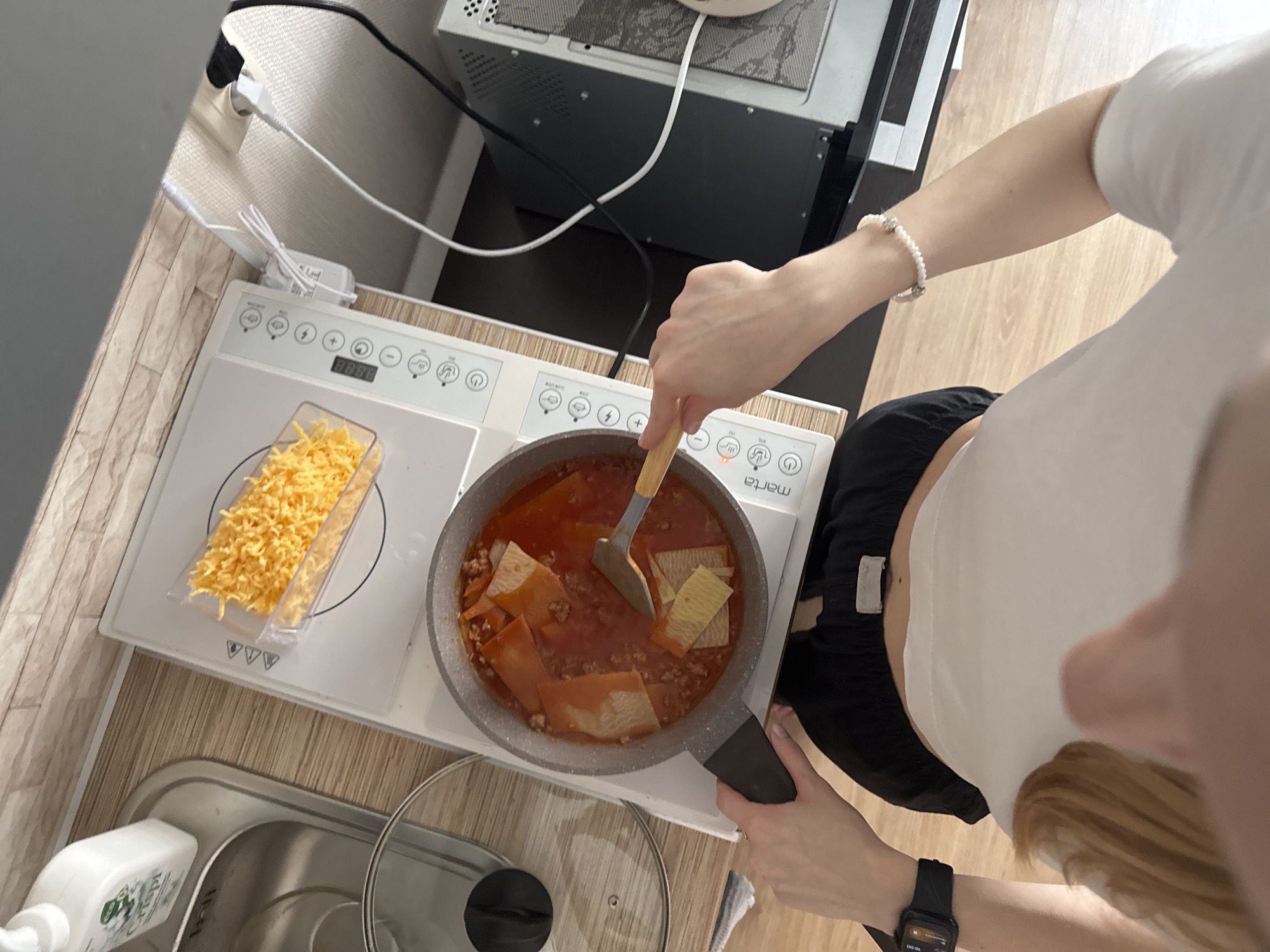 The image provides an overhead view of a person cooking in a compact kitchen or kitchenette. The subject is a light-skinned individual, seen from the chest down, wearing a white crop top and black shorts or pants. Their right arm, adorned with a pearl bracelet, is extended into the frame as they use a wooden spatula to stir a pan. A black smartwatch is visible on their left wrist.
The main activity is cooking. The person is stirring a mixture in a grey pan placed on a white portable induction cooktop. The pan contains a red sauce, ground meat, and flat, rectangular pieces of pasta, possibly for a lasagna-style dish. To the left of the cooktop, a clear plastic container holds a pile of shredded yellow cheese.
The cooktop is branded "Marta" and displays "888" on its digital interface, surrounded by various control icons for power, temperature, and timer settings. The kitchen area features a light-colored wooden or laminate countertop. To the left of the cooktop, a stainless steel sink is visible, with a white liquid soap dispenser beside it. A clear pot lid with a black handle rests near the sink. In the background, above the cooktop, is a dark grey appliance, possibly a microwave or oven, with a ventilation grate on its top surface. White power cables and plugs are visible along the wall, connected to outlets behind the appliances. The setting is indoors, likely a home kitchen in Perm, Russia, as suggested by the context and the commonality of the "Marta" brand in Eastern Europe. The scene is neutral, focusing on the practical act of preparing food.