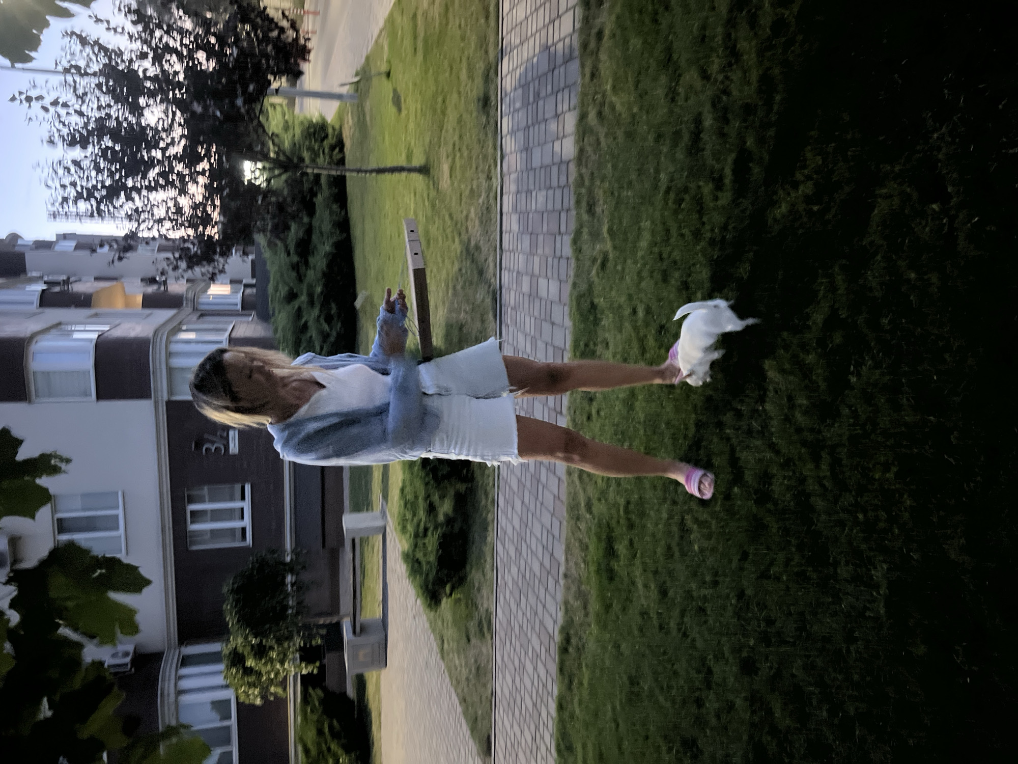 A woman in a light blue cardigan, white t-shirt, and white denim shorts stands on a grassy area. She is holding a pizza box in her hands. A small white dog is positioned near her feet, looking to the left. The woman is wearing pink slip-on sandals. Behind her, there is a paved walkway with square tiles and a low hedge. Further back, a multi-story apartment building with numerous windows and air conditioning units is visible. The building has a dark brick base and a lighter upper section, with a sign that reads "3/2". Trees and bushes are present in the background, and the sky appears to be dusk. The location is Kryzhanivka, Ukraine.