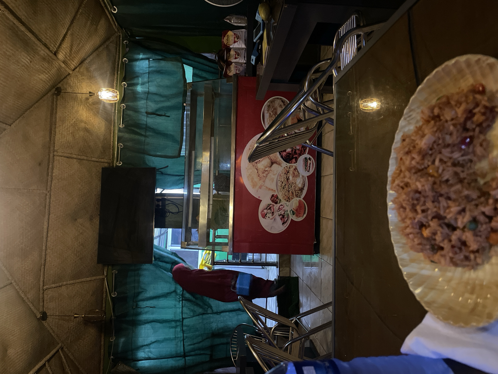 The image depicts an indoor dining area in what appears to be a restaurant in Hayin Banki, Nigeria. The scene is partially obscured by a plate of rice-based food in the foreground. There is a person with a red shirt near a refrigerator with food items on its side in the back. Green fabric covers some of the surroundings. Several metal chairs are visible. Lighting fixtures are present on the wall. The structure of the building is visible on the left side.