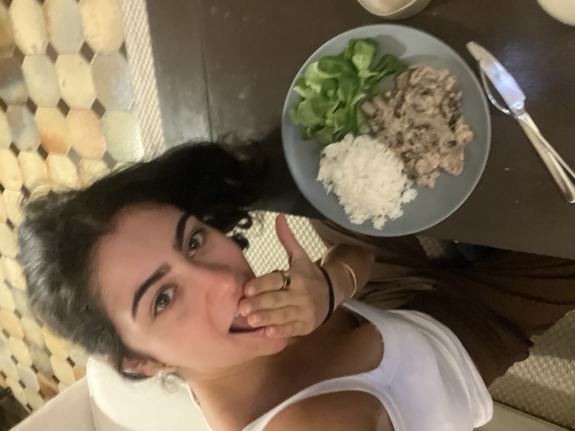 A person with dark hair and eyes, wearing a white tank top and jewelry on their left hand, is holding their hand to their mouth, seemingly in surprise or excitement. To the right of the person is a gray plate with three sections of food: rice, salad greens, and a creamy, meat-like mixture. A silver knife is placed on the right side of the plate on a dark brown surface. The background on the left shows a patterned wall, while the background on the right suggests a brown surface and possibly a carpeted area. This is likely a meal being enjoyed in a private residence. The location is unknown.