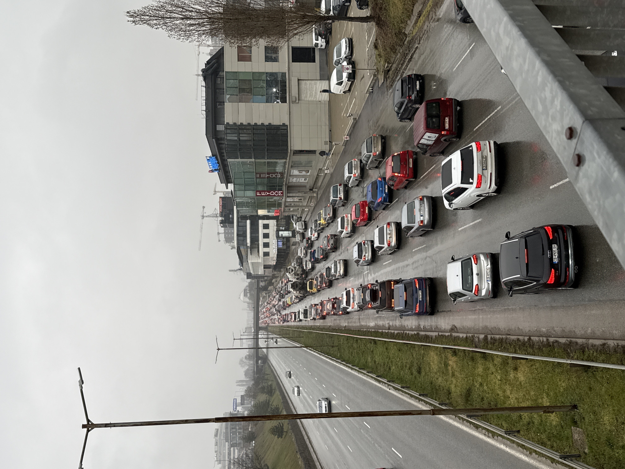 The image shows a traffic jam on a highway in Sofia, Bulgaria, on a cloudy day. The view is from an elevated position, likely a bridge. The highway is divided, with one side densely packed with cars stopped or moving slowly, and the other side with much less traffic. Cars are various colors, including red, white, black and blue. Alongside the highway, there is a strip of green grass, and beyond that, buildings are visible, including a shopping center with the brand "TEODOR" and possibly a construction site in the distance. The sky is overcast. A metal structure, possibly part of the bridge, is visible in the foreground. The cars in the right lane have license plates that are Bulgarian.