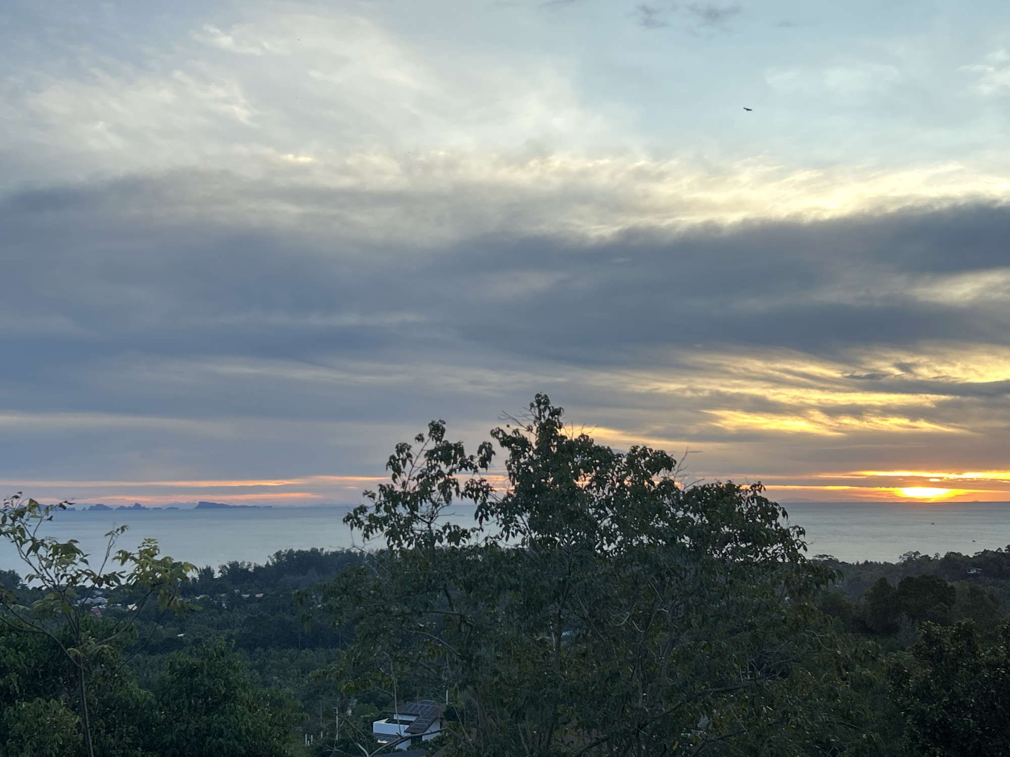 The image shows a scenic view at sunset in Baan Hin Kong, Thailand. The horizon features a body of water, likely the sea, with a scattering of islands in the distance. The sun is setting on the right side of the frame, casting a golden and orange glow across the water and sky. The sky is filled with layers of clouds, ranging in color from gray and blue to yellow and orange. A bird is visible in the sky. In the foreground, there are lush green trees. A building with a light-colored roof, potentially a residence, is partially visible among the trees. The scene appears peaceful and natural.