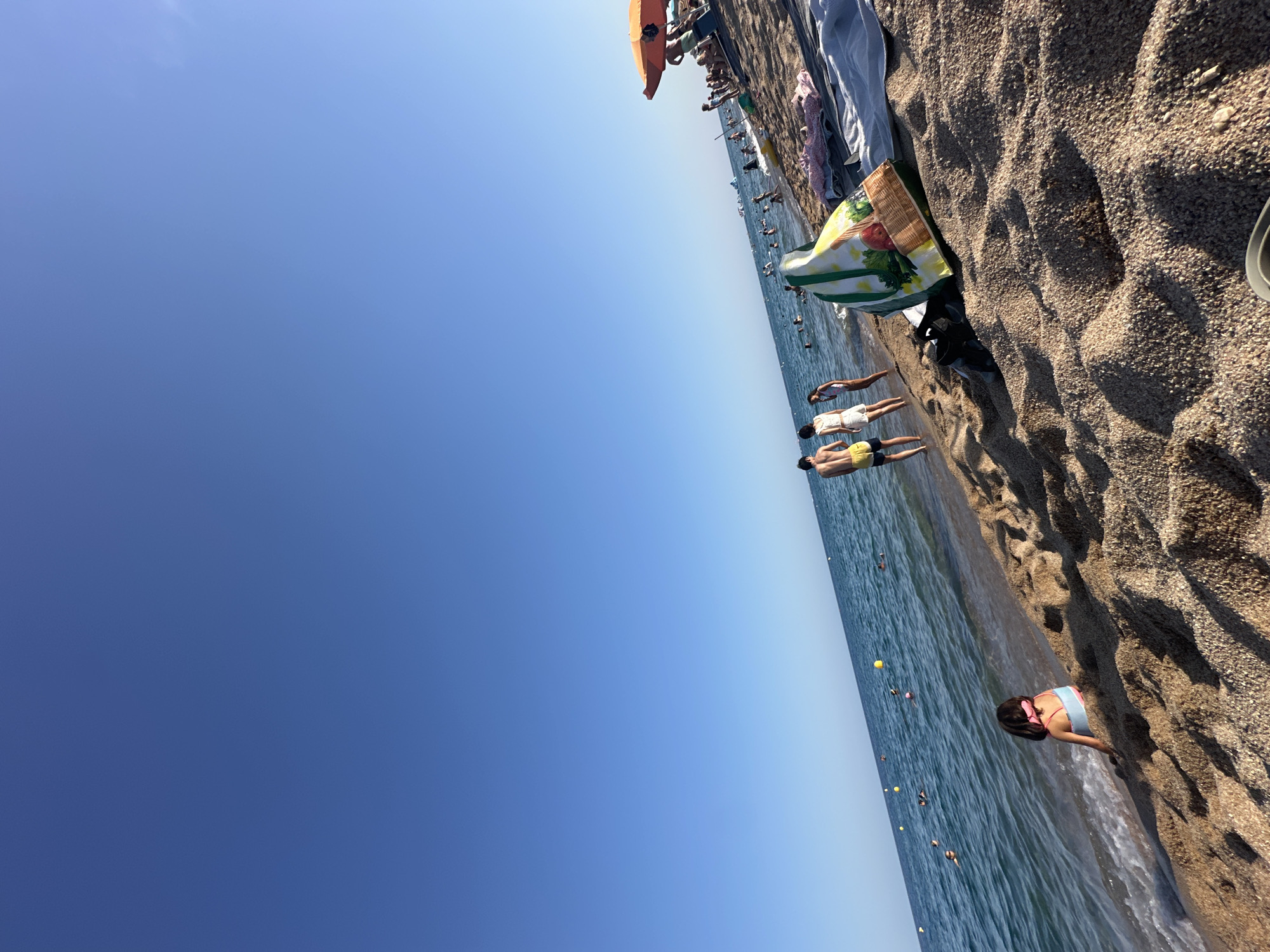 The image depicts a sunny day at the beach in Blanes, Spain. The clear blue sky stretches across the upper portion of the frame. The foreground is dominated by coarse, sandy beach material. In the middle ground, the tranquil blue sea meets the shore, with small waves lapping at the sand. Several people are visible in the water, some swimming and others standing near the shoreline. On the beach itself, a few individuals are present. To the left, a young girl in a blue and pink swimsuit sits facing the sea. Further to the right, a couple, a man in yellow shorts and a woman in a white top and shorts, stand at the edge of the water. A patterned beach bag rests on the sand in the foreground, and a folded towel is visible behind it. In the background, more beachgoers and some boats are scattered across the water. An orange umbrella is set up on the beach to the far right, providing shade. The overall scene conveys a relaxed summer atmosphere.