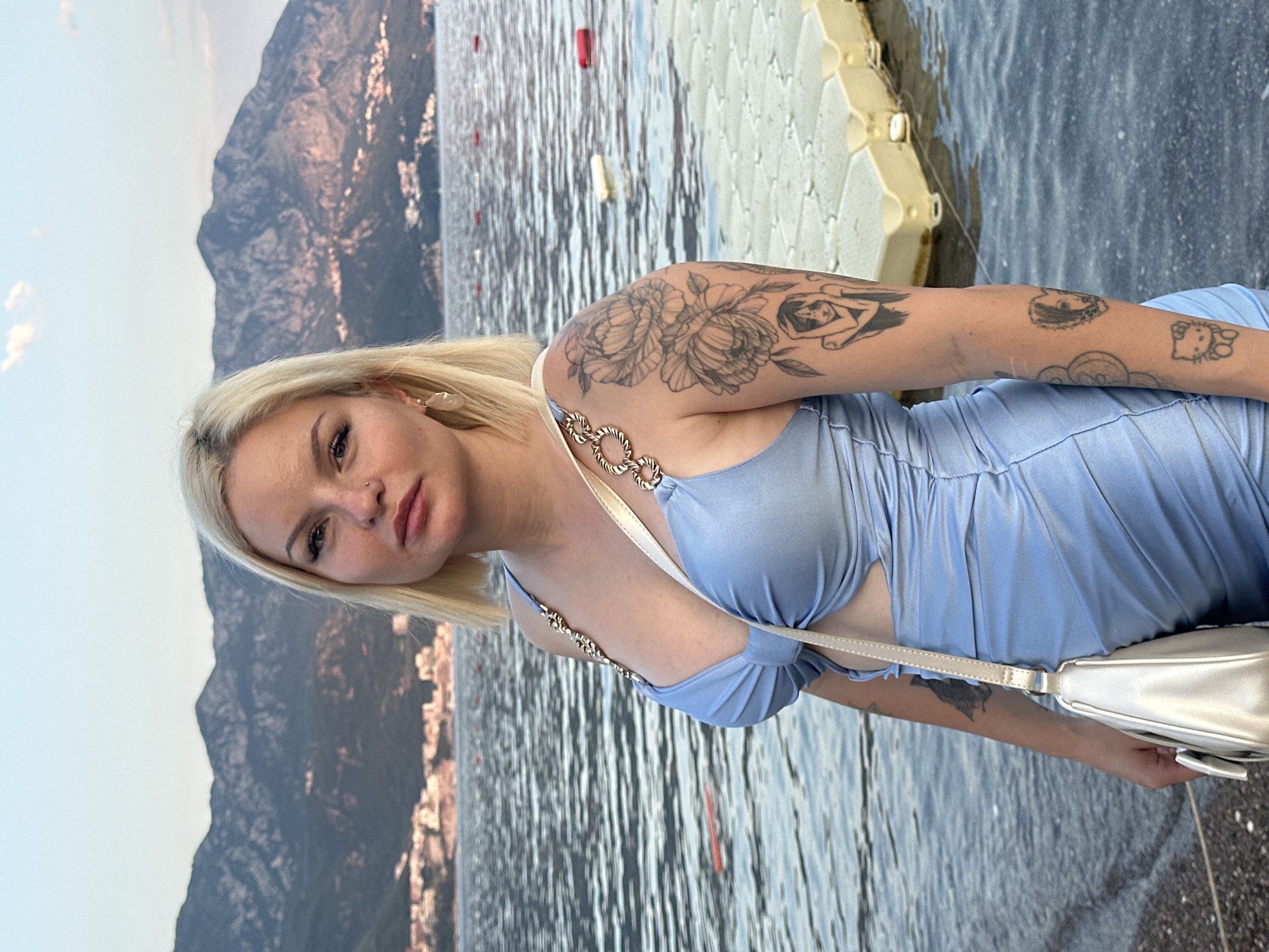 A young woman with blonde hair is posing for a photo in an outdoor setting. She is wearing a light blue, ruched dress with chain detailing on the straps and a cutout at the waist. She has a small handbag slung over her shoulder. Her left arm, visible in the foreground, is adorned with several tattoos. The woman has a neutral expression and is looking towards the camera.
The background of the image shows a body of water, likely the sea, with ripples on the surface. In the distance, a large, mountainous landscape is visible, with some buildings clustered at the base of the mountains. A light-colored, modular floating dock is also present in the water to the right of the woman. The lighting suggests it is daytime, possibly late afternoon or early evening, given the soft light and shadows. The overall atmosphere appears to be relaxed and scenic. The location cues, specifically the mountains and the coastline, along with the provided context, strongly suggest this scene is set in Boreti, Montenegro. There is no visible text in the image.