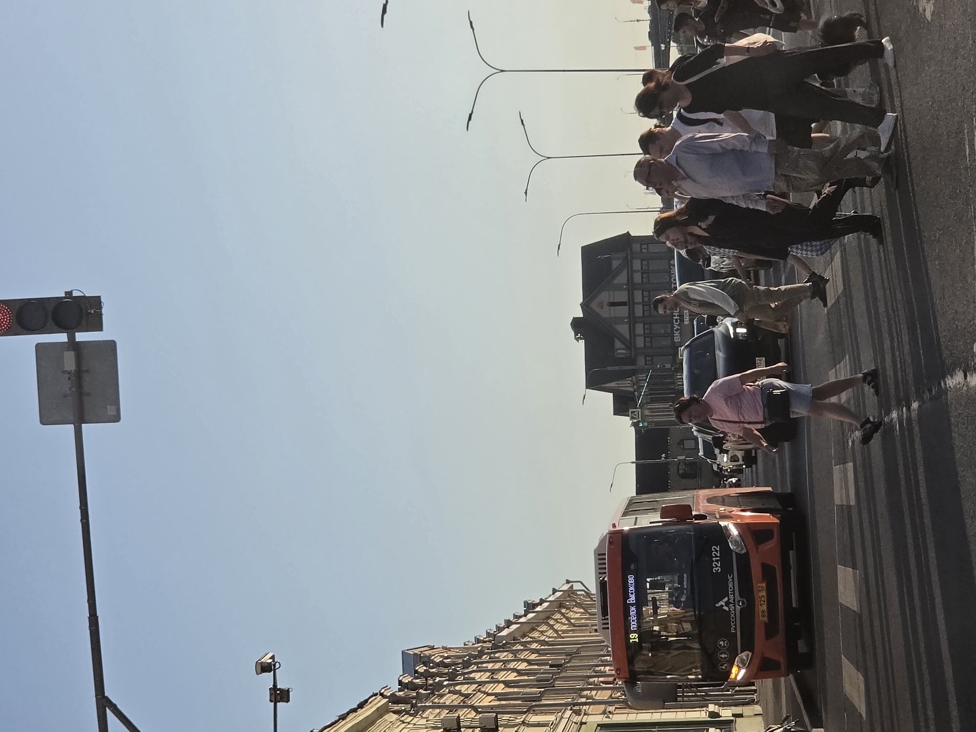 In this street-level, medium-shot image captured in Nizhny Novgorod, Russia, a group of pedestrians crosses a well-lit, paved intersection with clear pedestrian markings. The sun, low in the sky, casts long shadows across the road, suggesting it is either early morning or late afternoon.
The primary subject in the foreground is a woman in a pink t-shirt and denim shorts, walking from right to left, her gaze directed forward. To her right, a dark-colored SUV is partially visible, and a second pedestrian, a woman in a black outfit, is seen behind her, also walking from right to left.
Further back, a group of individuals is crossing from left to right. Among them, a man in a light blue button-down shirt and camouflage pants is prominent, alongside a woman in a black jacket and a young person in a white t-shirt. Another man in a light-colored jacket and tan pants is positioned to the left of this group.
Dominating the left side of the frame is the front of an orange bus, displaying the route number "19" and Cyrillic text that translates to "poselok Vysokovo." The bus has its headlights illuminated. The bus's license plate reads "BB 125 52."
In the background, a building with a distinct architectural style, featuring multiple windows and a mansard roof, is visible. Traffic lights are present, with one displaying a red signal at the top of the frame. Streetlights with multiple lamps line the street, and a traffic sign is mounted on a pole. The overall environment appears to be an urban setting with buildings lining the street and clear blue sky above.