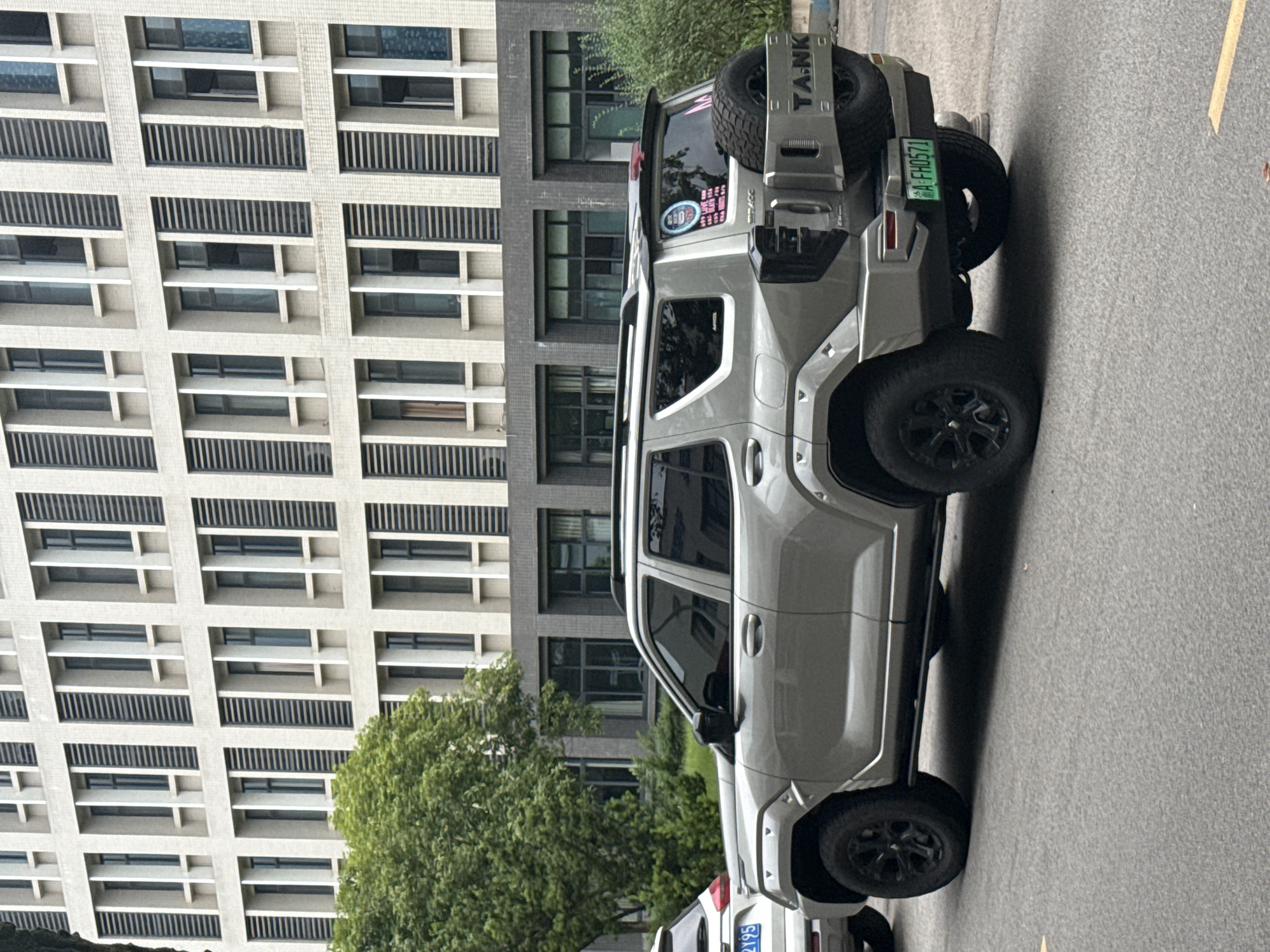 A silver "TANK" SUV is parked on a street in Jiangjin, China, in front of a large building with multiple windows. A green license plate reading "渝A FH0571" is visible on the rear of the vehicle. Another vehicle with the license plate "渝A 21Y95" is visible in the lower left corner. A tree partially obscures the view. The street appears to be paved with asphalt.
