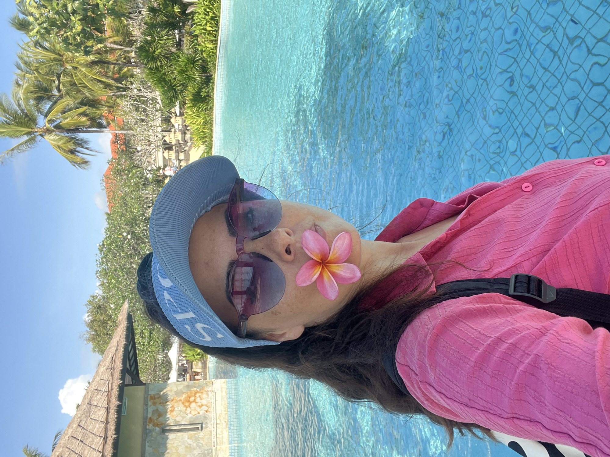 A woman with long dark hair wears a pink button-up shirt, a blue visor, and sunglasses. She holds a pink and yellow flower in her mouth while standing by a bright blue swimming pool. Palm trees and lush green foliage are visible in the background, along with a thatched-roof building and a resort structure. The scene is set in Kuta Selatan, Indonesia, a location known for its tropical atmosphere and luxurious resorts.