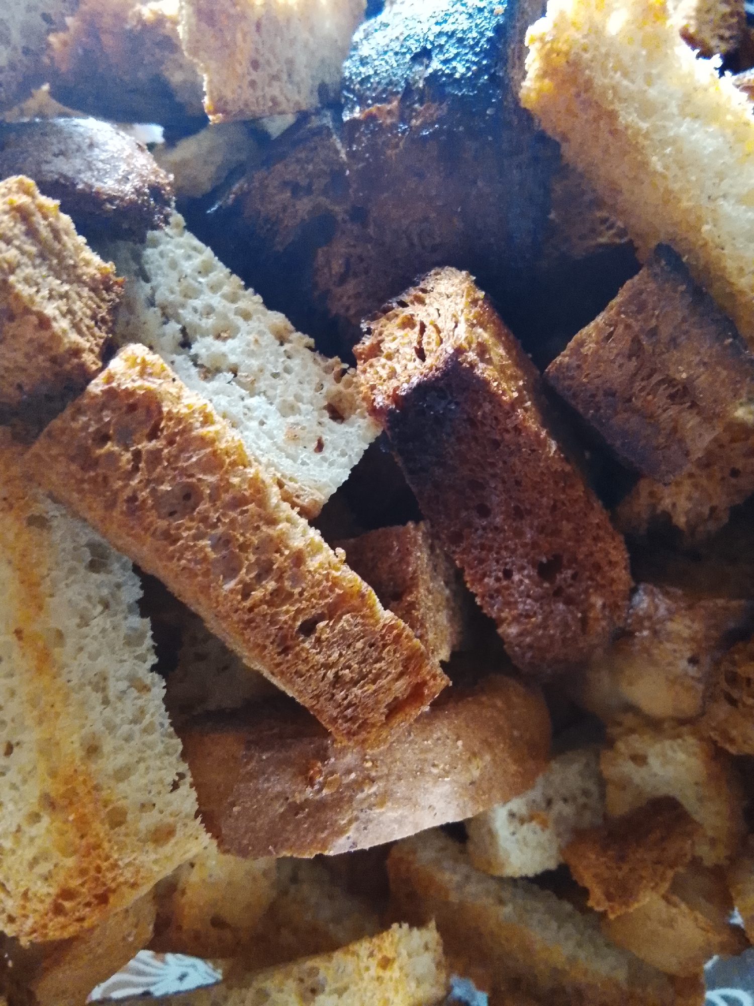 The image displays a close-up, overhead view of a pile of dry bread pieces, commonly known as croutons or rusk. The bread pieces vary in color, ranging from light beige to dark brown, indicating different types of bread or degrees of toasting. The texture of the bread is rough and porous, with visible air pockets. The lighting creates highlights and shadows, adding depth to the scene. The background is dark and indistinct, focusing all attention on the bread. There are no people, animals, or discernible environmental features present in the image. The composition suggests a food-related context, possibly a kitchen or pantry. The image does not contain any elements that would allow for its localization to Almaty, Kazakhstan, or any other specific city.