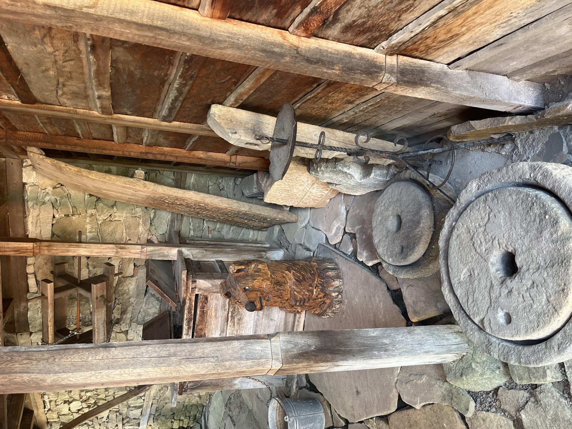 The image depicts the interior of a rustic wooden structure, likely a historical exhibit or outbuilding, situated in the town of Zheravna, Bulgaria. The floor is paved with irregular flat stones, and the walls are constructed from rough-hewn stone. Dominating the foreground are two large, circular millstones, one partially visible in the lower center and another positioned behind it. To the left, a weathered wooden post supports the structure, with a metal bucket hanging from a chain nearby. Further back, a carved wooden bear sculpture sits on the stone floor. Various other wooden elements, including beams, planks, and a wooden structure with chains and gears, are visible throughout the space, suggesting it may have been a workshop or storage area. A wrought-iron candle holder stands on the right side of the image, leaning against the wooden wall. The overall impression is one of age and tradition.