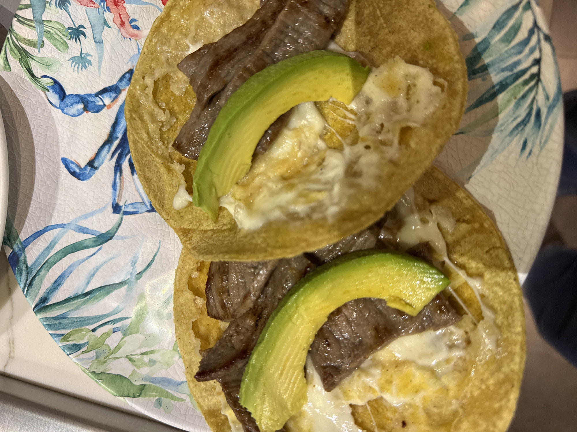 Two tacos filled with meat, cheese, and avocado slices are arranged on a decorative plate with floral and leaf patterns.
