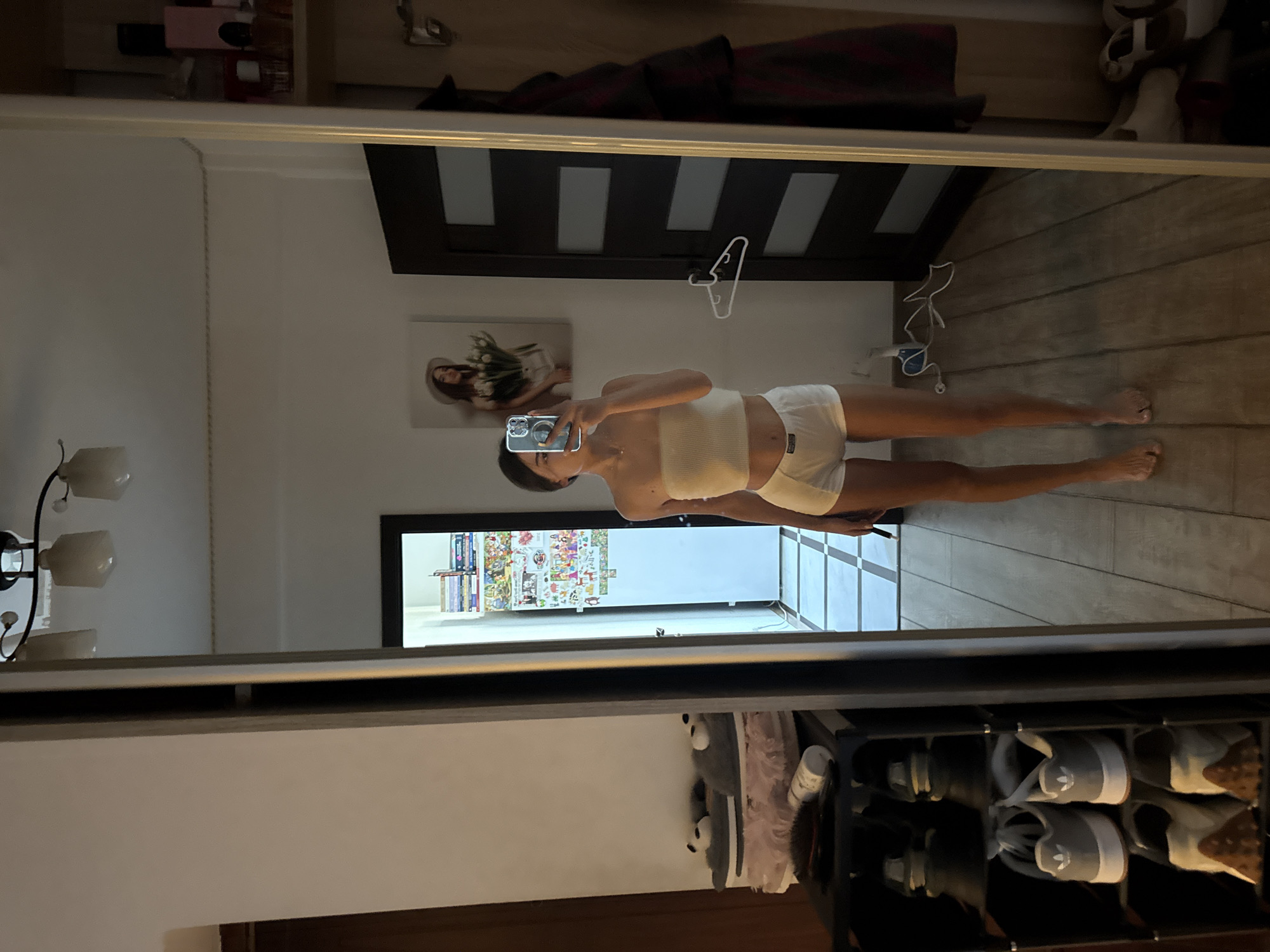 A young woman is standing in a room, taking a mirror selfie. She is wearing a white bandeau top and white shorts. She has dark hair pulled back and is holding a phone in her right hand.
The room appears to be a bedroom or dressing area. To the left of the woman, there is a tall mirror reflecting the scene. In the mirror's reflection, we can see a shoe rack filled with various shoes, including sneakers and slippers. Above the shoe rack, there are some soft slippers and a hairbrush.
To the right of the woman, there is a dark door with horizontal panels, and next to it, a wardrobe with clothing hanging inside. On the wall above the door, there is a framed picture of a woman holding tulips. Further to the right, in the mirror's reflection, there are shelves with perfume bottles.
The floor is covered with wooden planks, and there are checkered tiles in the background, suggesting a hallway or an adjacent room. A white clothes hanger is hanging on the door. The lighting in the room is dim, with some artificial lights visible on the ceiling.
The image does not provide any specific clues about the location within the city of Chișinău, Moldova.