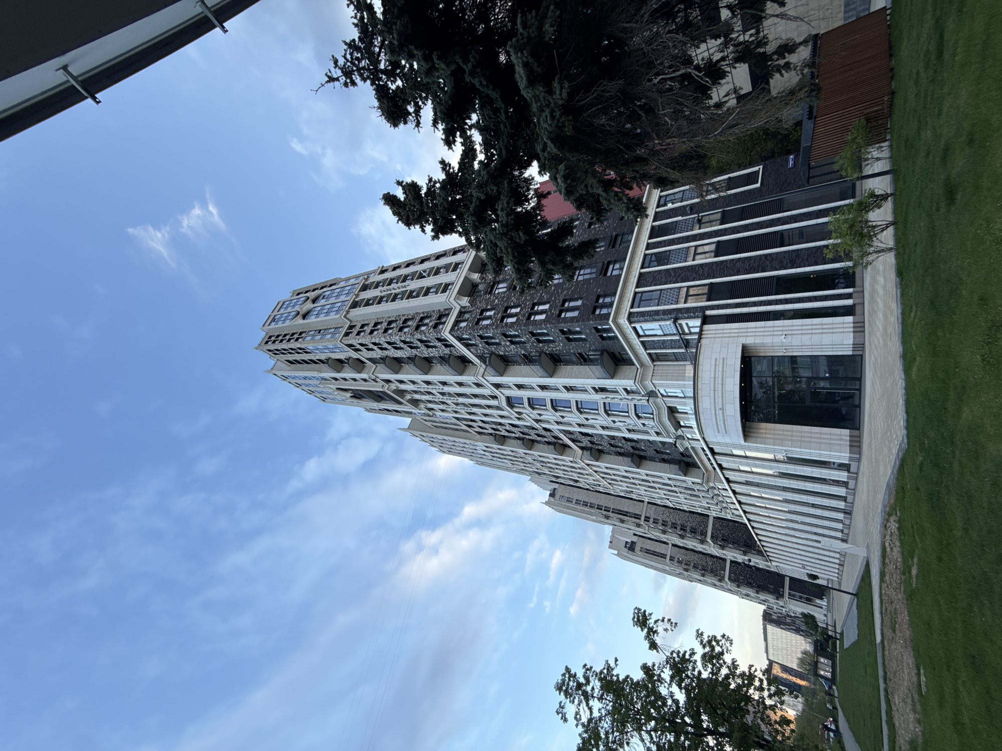 The image depicts a tall residential building in Khimki, Russia, viewed from a low angle. The building has a modern design with alternating sections of light and dark facade elements. Trees are visible in the foreground, partially obscuring the lower portion of the building. The sky is blue with scattered clouds. A patch of green grass is visible along the building's base, suggesting a landscaped area.