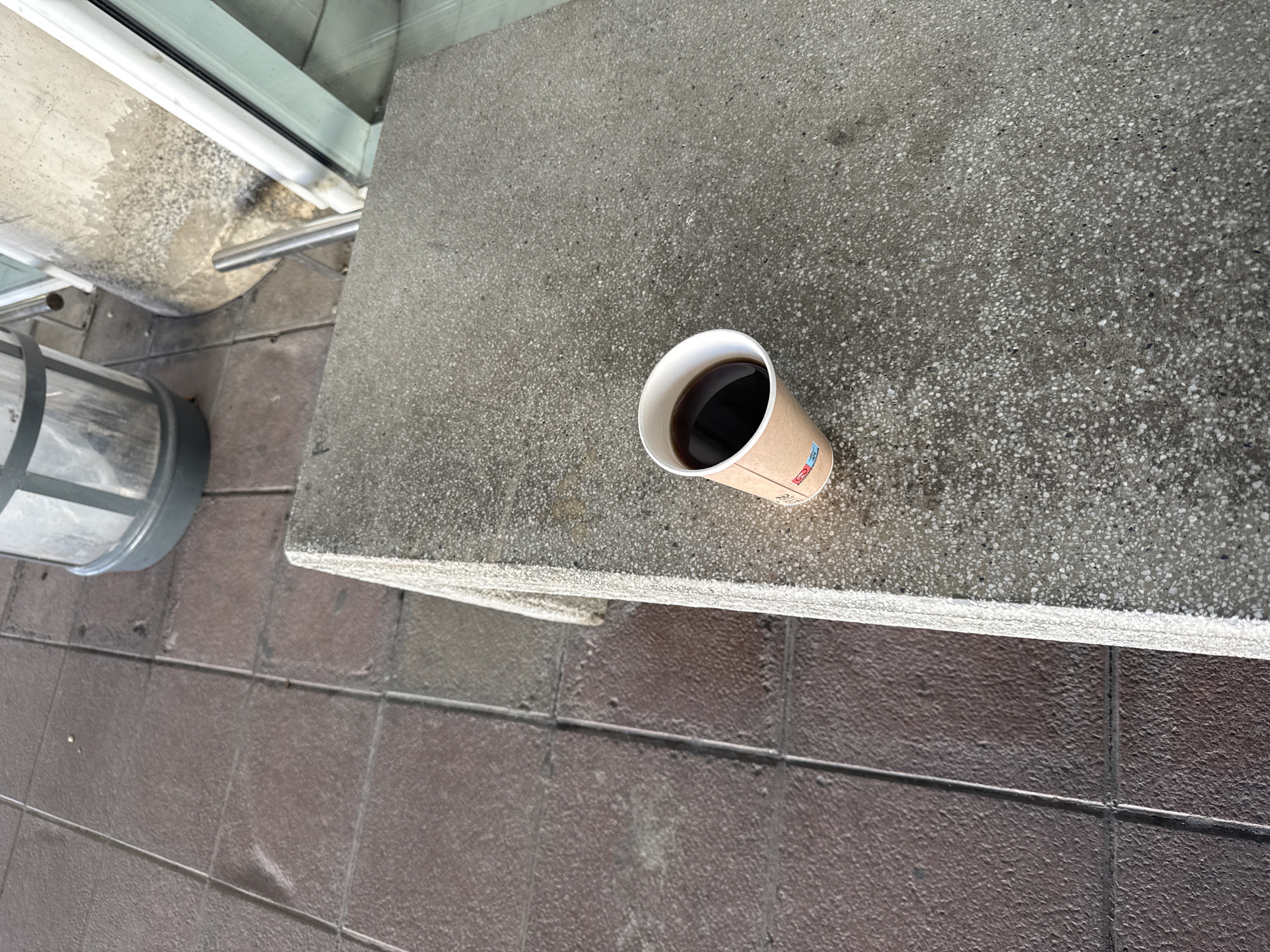 The image shows a cup of coffee resting on a concrete surface. A metal trash can is visible to the left, and a tiled surface is at the bottom left. Part of a window is at the top left.
