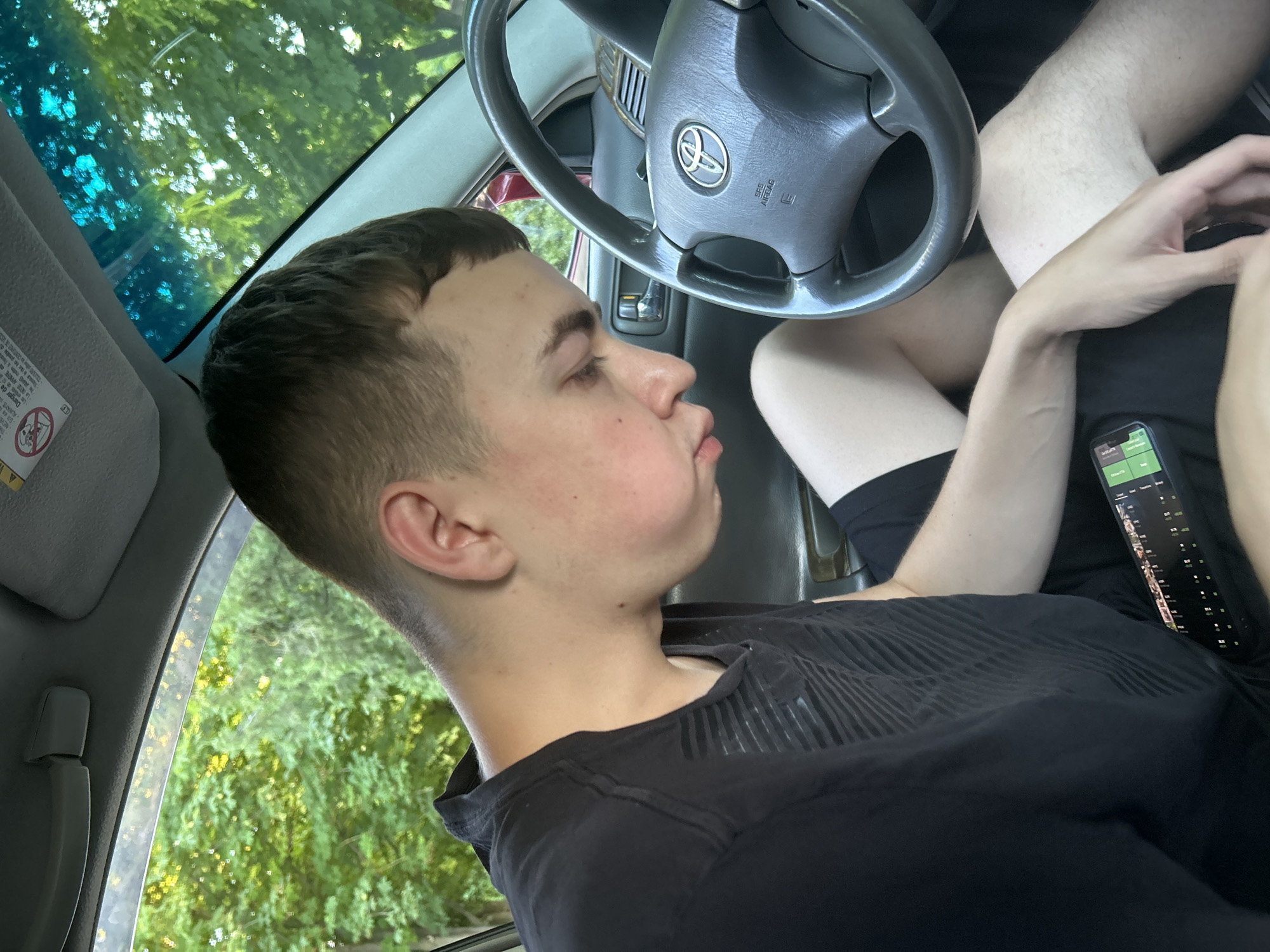 A young man with short brown hair and fair skin is sitting in the driver's seat of a car, looking down. He is wearing a black t-shirt and black shorts. His left hand rests on his lap, holding a smartphone. The car's steering wheel is visible, with a Toyota logo in the center. Through the car windows, lush green trees and foliage can be seen, suggesting an outdoor setting, possibly in a rural area or a park, in the vicinity of Ivanivka, Ukraine. The lighting indicates it is daytime, with sunlight filtering through the trees.
