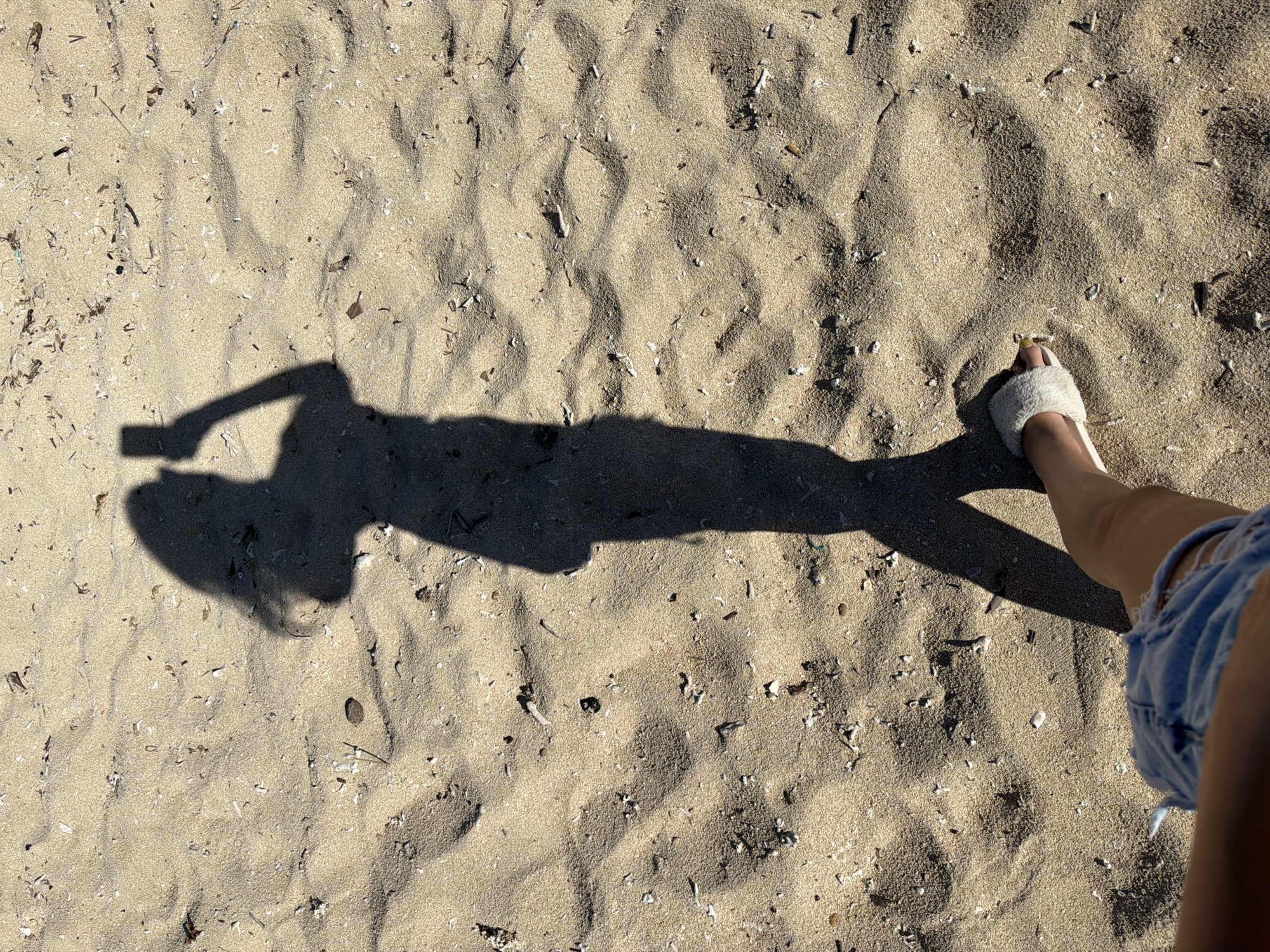 The image shows a shadow of a person holding a mobile phone, standing on a sandy beach. The person's legs and feet are visible in the foreground, wearing denim shorts and a fluffy slipper. The sand is light brown and scattered with small shells and debris. The scene suggests a sunny day, likely at a beach in Kute, Indonesia. The shadows indicate the sun is at an angle, suggesting it is either morning or late afternoon. The overall impression is a candid shot of someone taking a selfie or a picture of their surroundings.