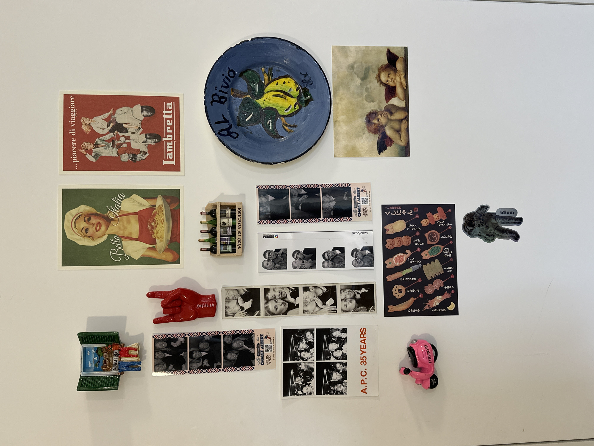 The image shows a collection of items attached to a white surface, likely a refrigerator door. The items are various souvenirs, photographs, and decorations. These include a red postcard featuring a vintage scooter with the word "Lambretta," a postcard with the text "...piacere di viaggiare" above the scooter, and a postcard of a woman holding a plate of pasta labelled "Bella Italia." Other items include a painted plate with blue edges, a postcard of two cherubs, a small wooden rack of miniature wine bottles labelled "VINI IN TOSCANA," several strips of black-and-white photo booth pictures, a red hand-shaped magnet with the word "SICILIA," a green window magnet labelled "TAORM INA", a postcard reading "A.P.C. 35 YEARS", a pink scooter magnet labelled "FIRENZE", a small menu-like sticker with cartoonish Japanese food items, and a magnet of an astronaut figure with the word "idiom" on it. The arrangement of the items is somewhat random, suggesting a collection accumulated over time.