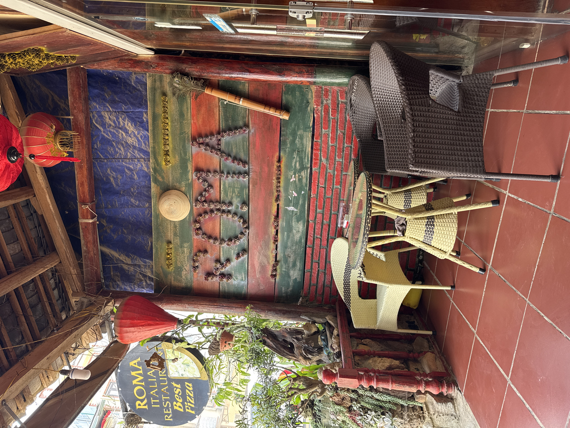 The image depicts an outdoor seating area of the Roma Italian Restaurant in Đồng Văn Commune, Vietnam. The restaurant's sign is visible, advertising "Best Pizza". The seating area features rattan chairs around a glass-topped table, placed on red tiled flooring. The backdrop is a wall decorated with painted wooden planks in red, green, and blue, adorned with pine cones arranged to spell "ROMA", a bamboo flute, and hanging decorations including lanterns and straw hats. Vegetation spills over the edge of a planter near the bottom of the frame.