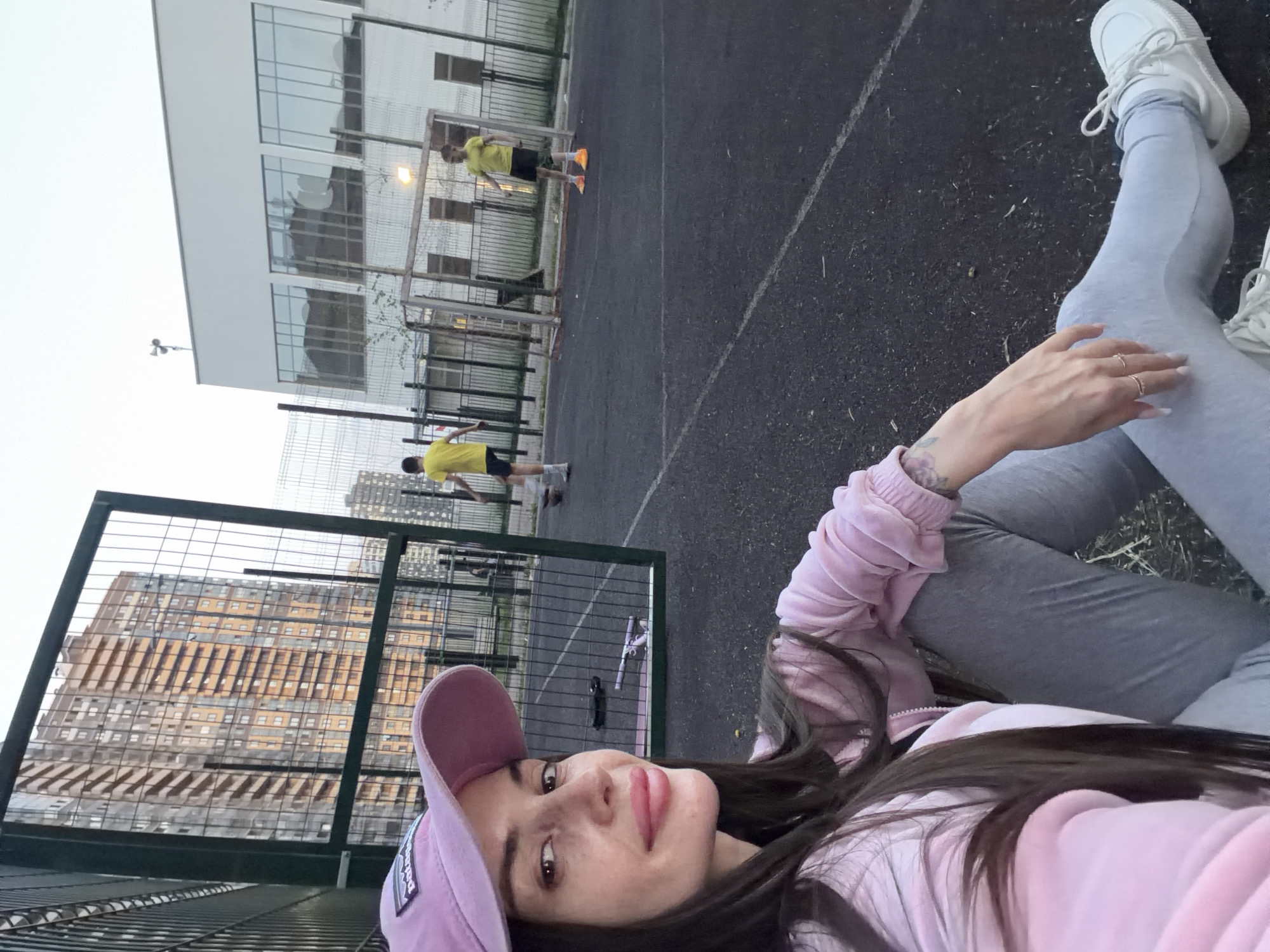 A selfie depicts a woman in a pink jacket and lilac baseball cap lying on a gray surface, possibly a playground or court, given the black lines visible. She also wears grey leggings and white sneakers. In the background, two people wearing yellow shirts and black shorts are playing or practicing soccer, as evidenced by the soccer goal with orange cones. The location is an urban area of St. Petersburg, Russia, identifiable by the presence of characteristic buildings behind the soccer court with a metal fence.